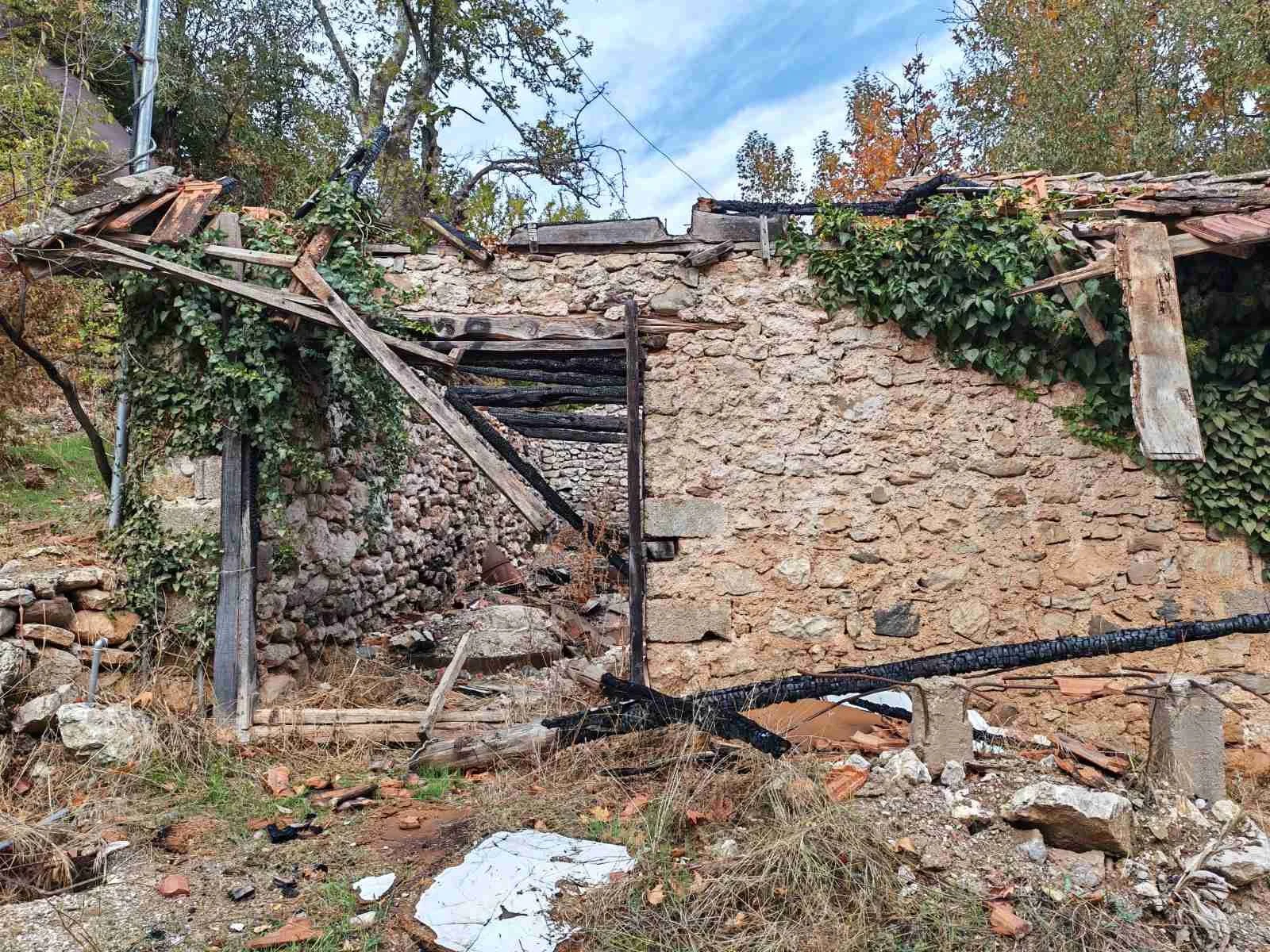 Yıkılmış ve hasar görmüş taş duvar ve ahşap yapının kalıntıları, etrafında yanmış veya hasar görmüş malzemeler ve dağınık doğa ortamı.