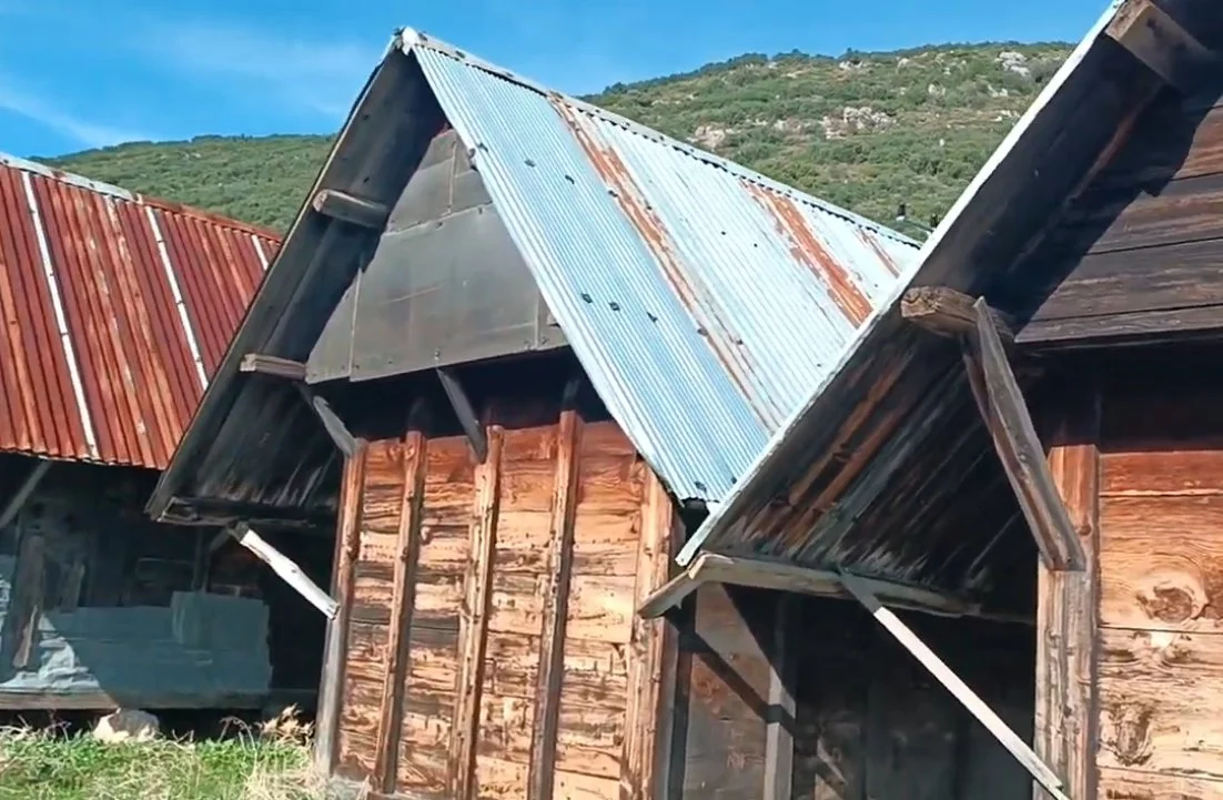 Eskimiş ve paslı metal çatılı ahşap evler, arka planda yeşil tepeler ve mavi gökyüzü.