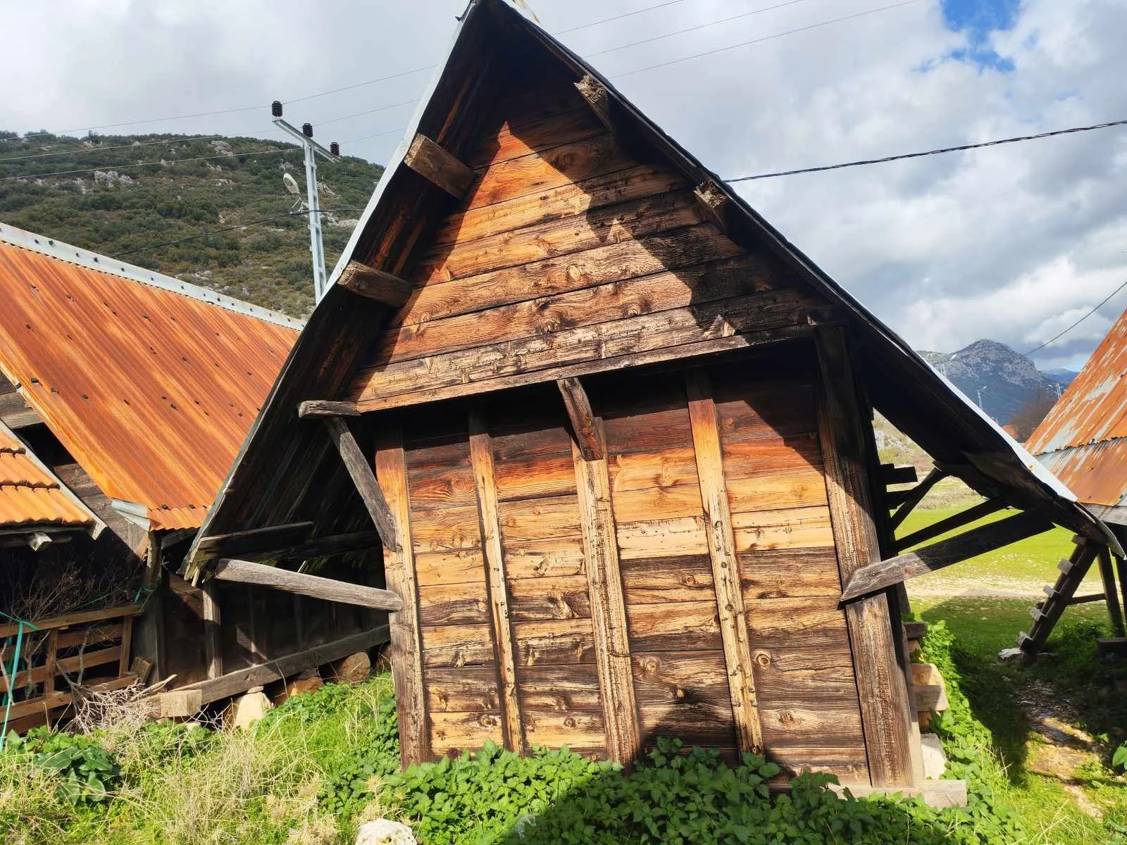 Ahşap kulübe ve çatısı, çevresinde yeşilliklerle çevrili, dağlar ve gökyüzü görünmekte.