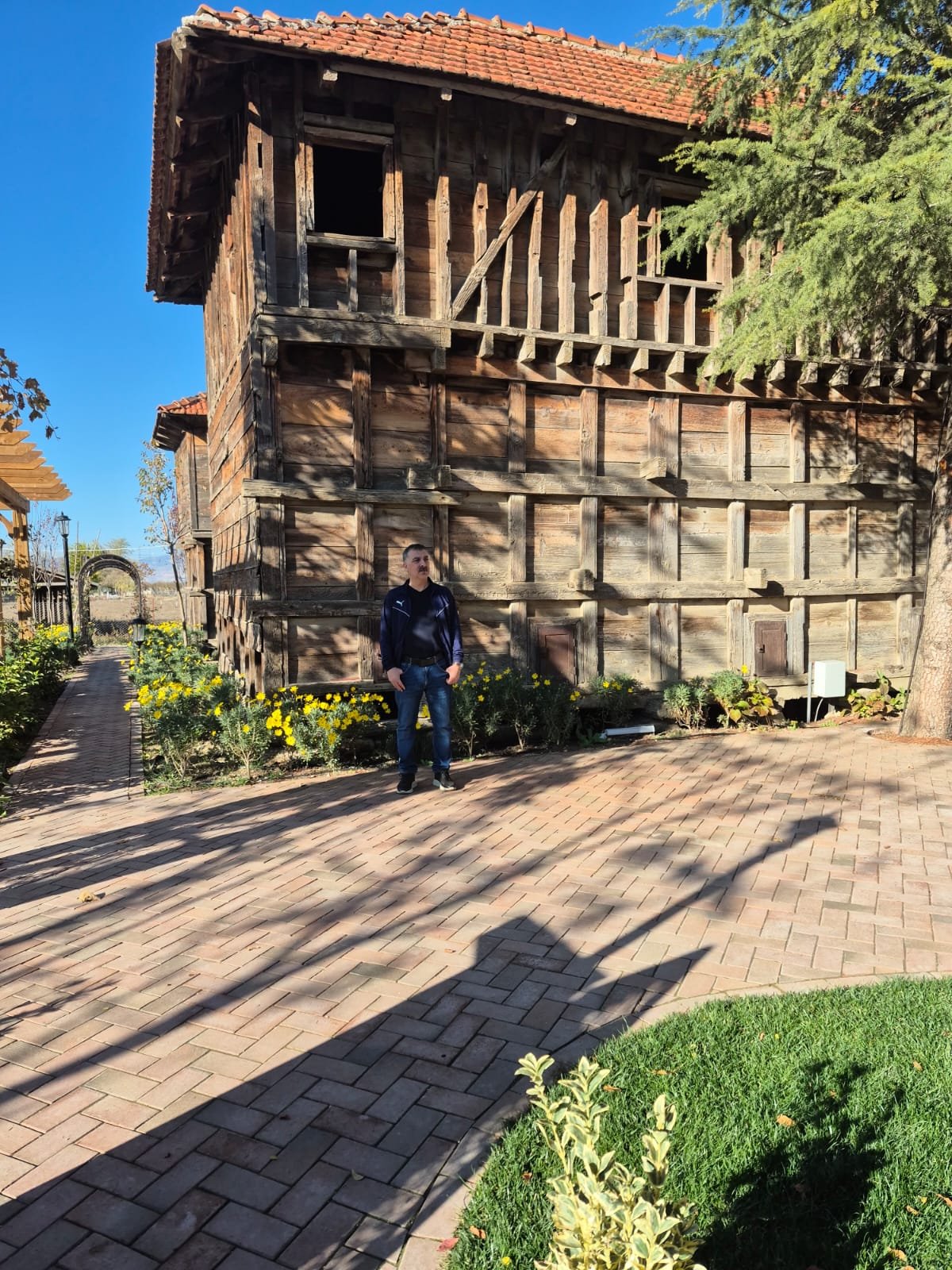 İnsan, eski ahşap bir binanın önünde duruyor. Binanın üst katı açık ve yapım sırasında gibi görünüyor. Çevrede çiçekler ve ağaçlar var, gölgenin olduğu alanlar zeminde görebiliyor.