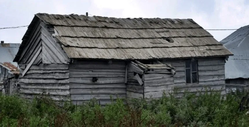Eski, ahşap ve çatısı hasar görmüş bir ev, çevresinde çalılık ve diğer eski yapılar var.