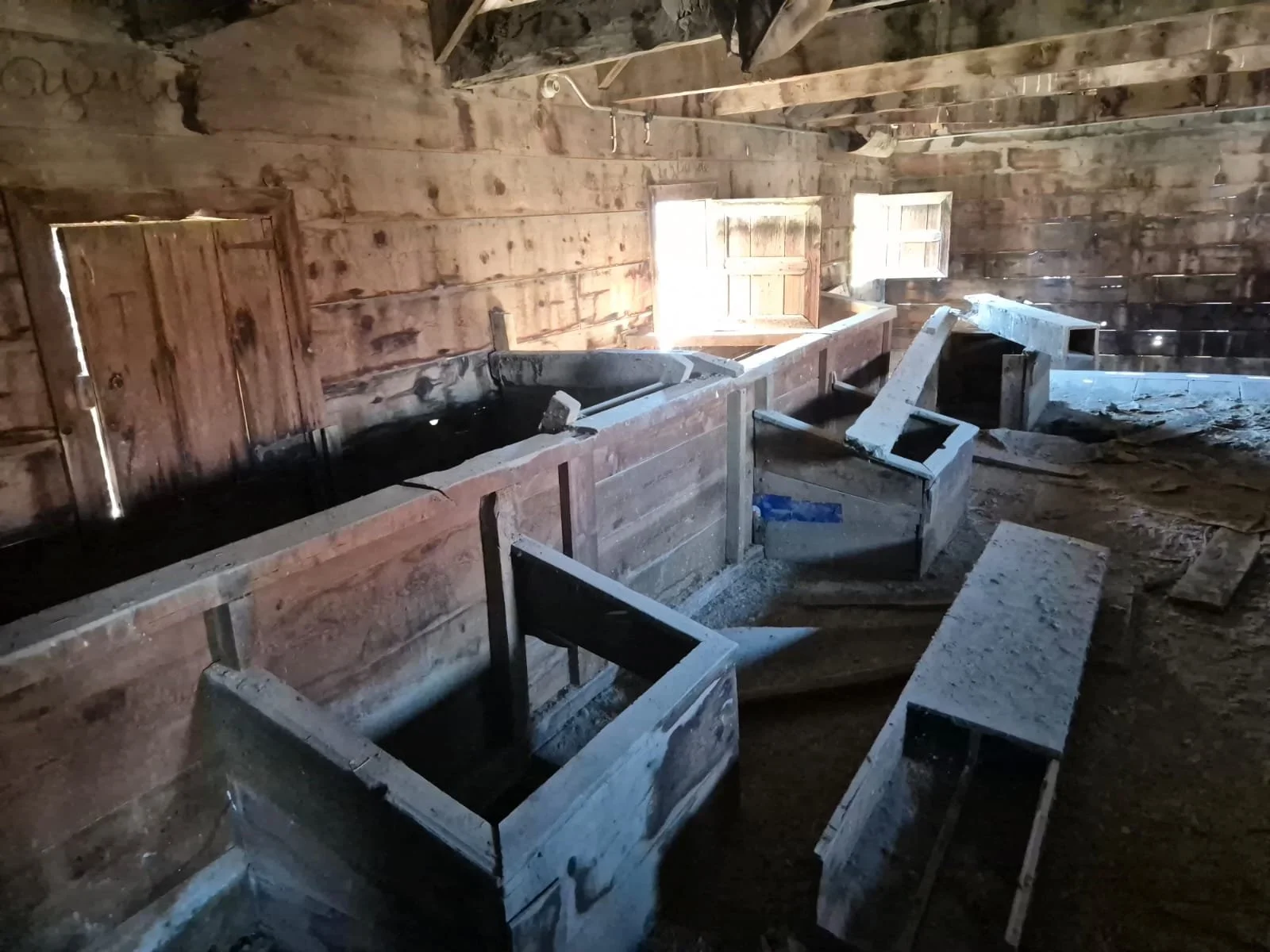 Ahşap duvarlarla ve pencerelerle eski bir yapıda, su banyosu ve çeşitli ahşap yapılar var, bazı eşyalar ve yüzeyler toz ve pislikle kaplı.