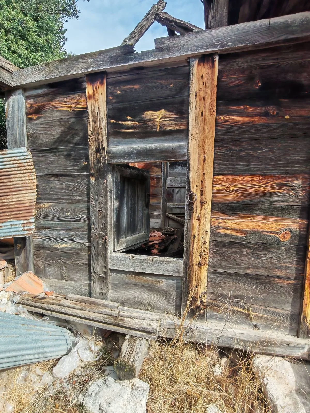 Eski, yıkılmak üzere olan ahşap bir bina, penceresi kırık ve kapısı açık, etrafta kuru otlar ve yamuk taşlar var.