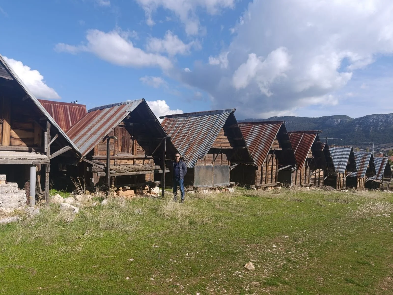 Eski ahşap ve metal çatılı yapılar sıralanmış, bir kişi ön planda duruyor, arka planda da dağlar ve gökyüzü var.