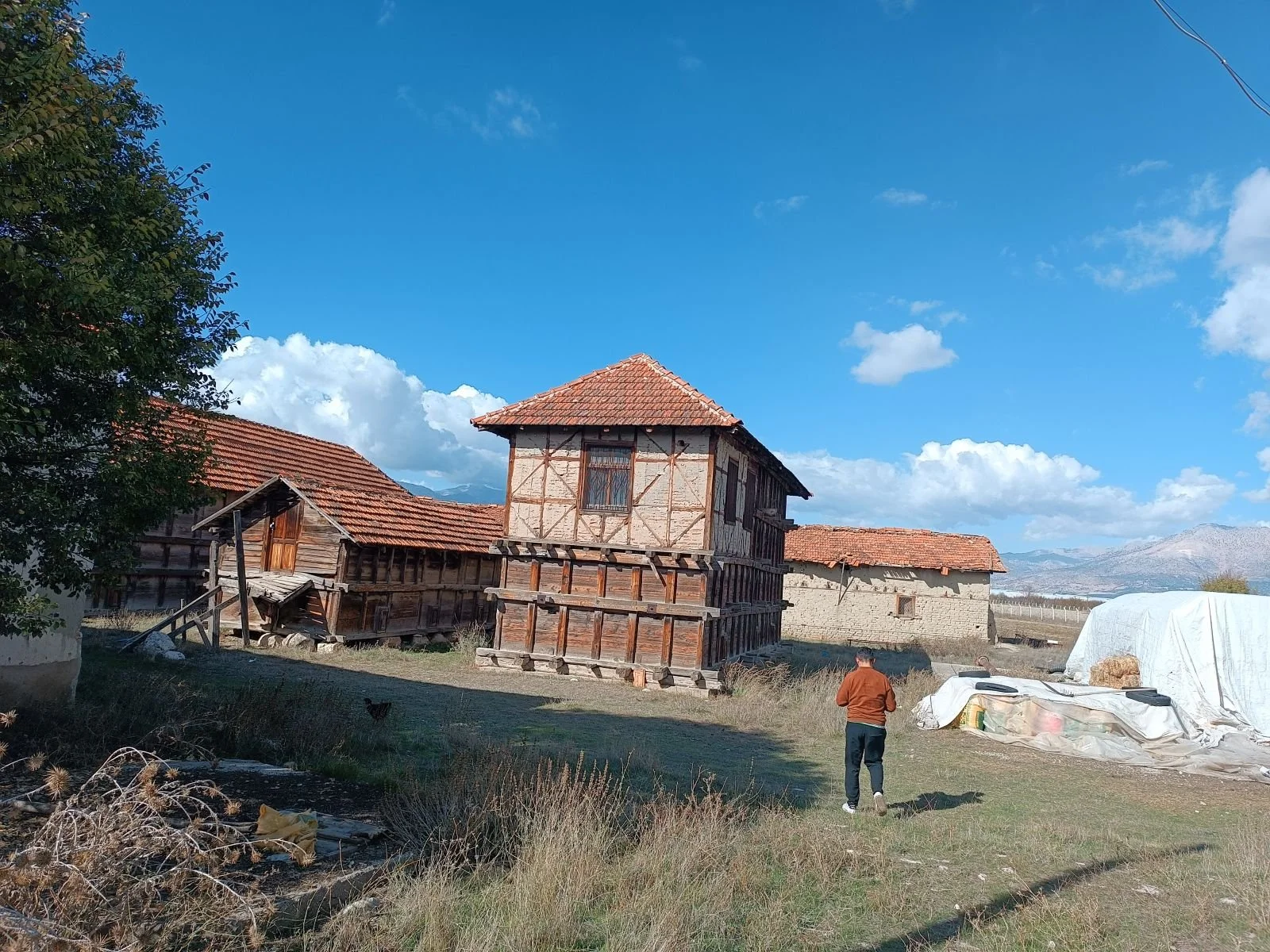 Eski taş ve ahşap evler, açık mavi gökyüzü ve dağlar ile kırsal bir alanda yer alıyor. Bir kişi uzaklarda duruyor.