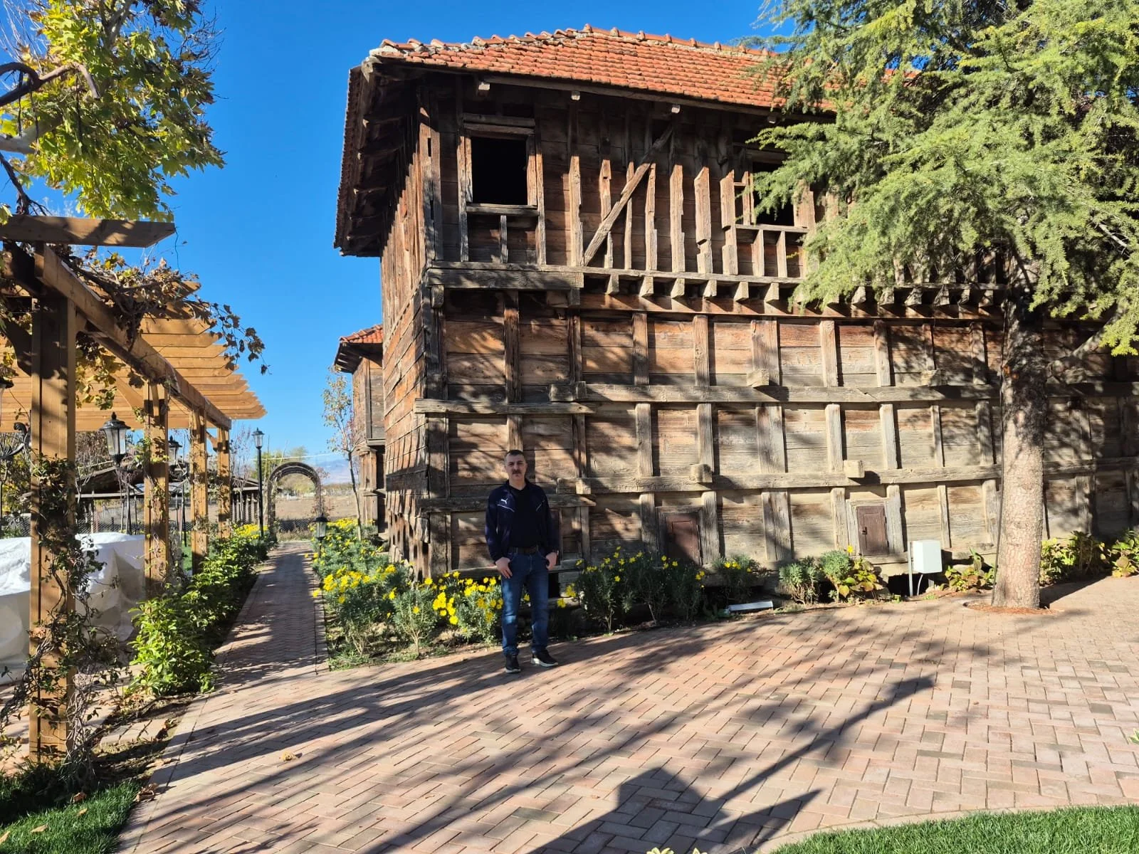 Ahşap tarihi ev ve bahçe. Bir adam önünde duruyor, gün batımı ve gölgeler var.