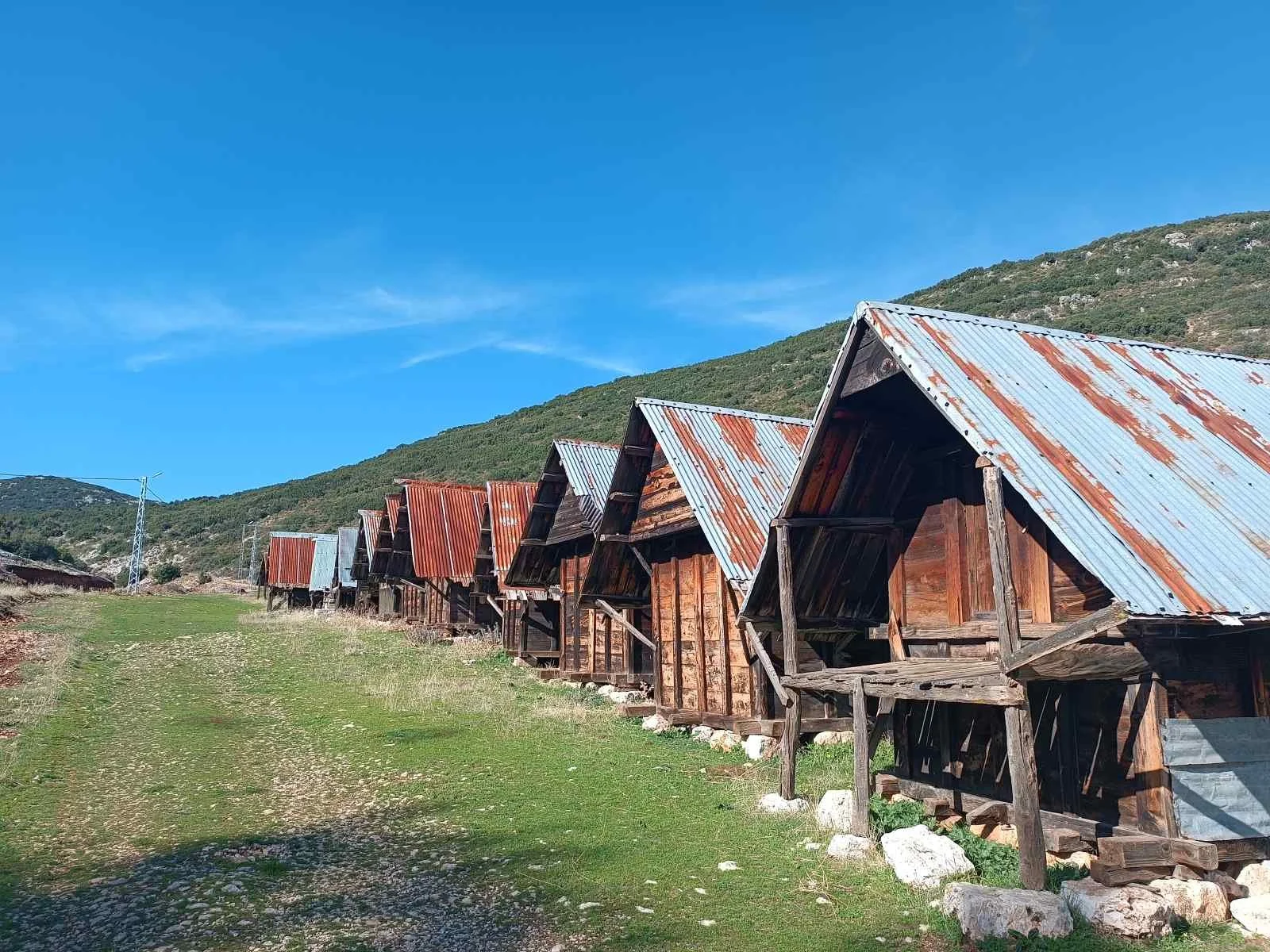 Dağ manzarası önünde eski ahşap kulübeler, çatılar metal levha ve ahşap ile kaplı, gökyüzü açık ve bulutsuz, yeşil alan üzerinde toprak ve taş yollar.