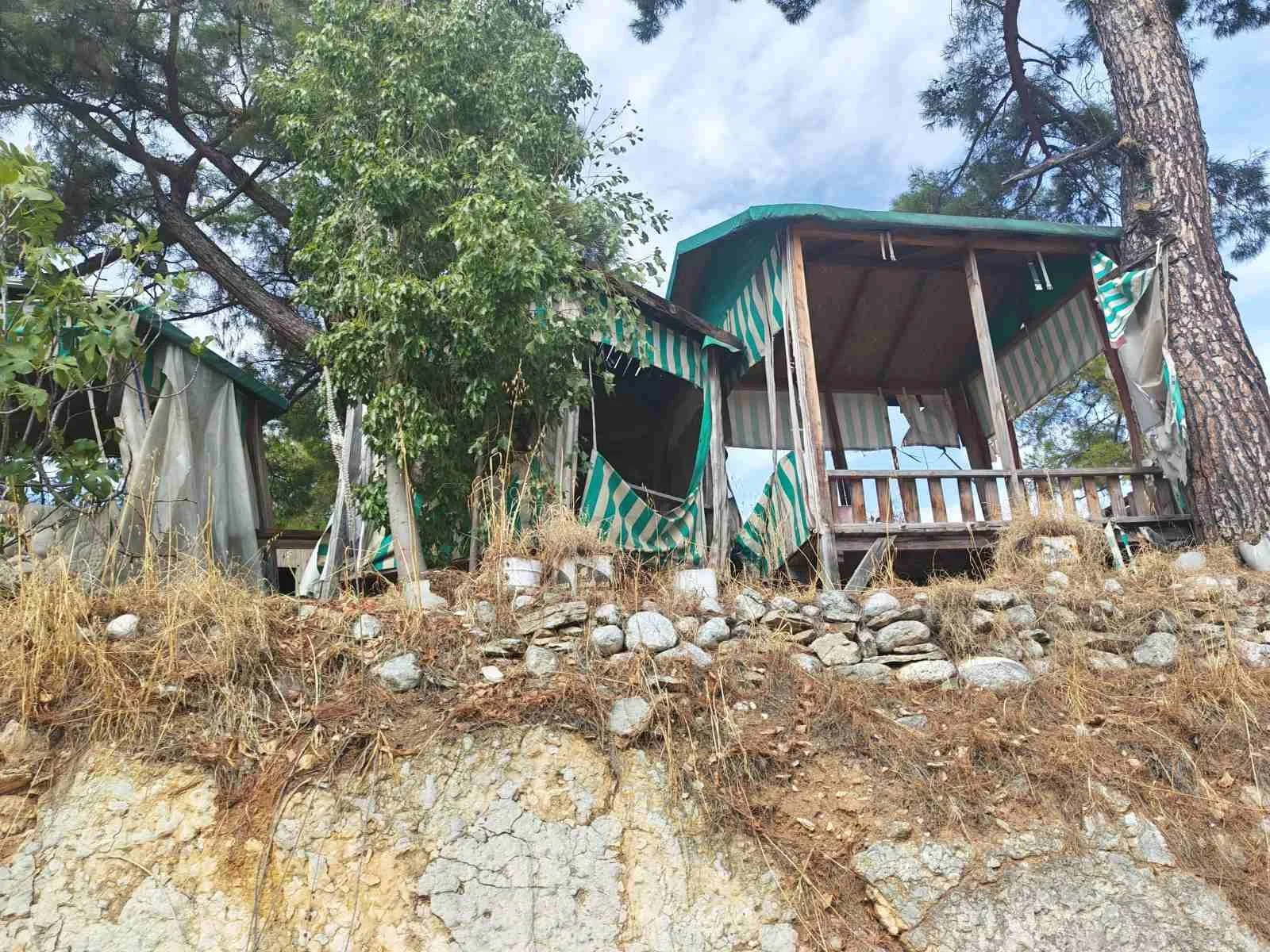 Eski ve bakımsız ahşap yapılar, çimenler, taşlar ve ağaçlar bulunan dağlık bir alan.