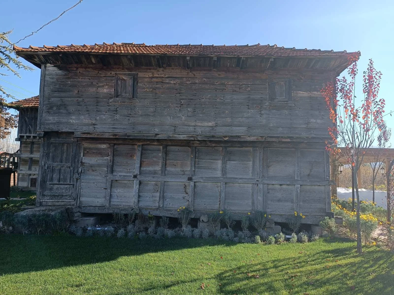 Ahşap eski bir ev, çevresinde yeşil çim ve bazı ağaçlar bulunuyor.