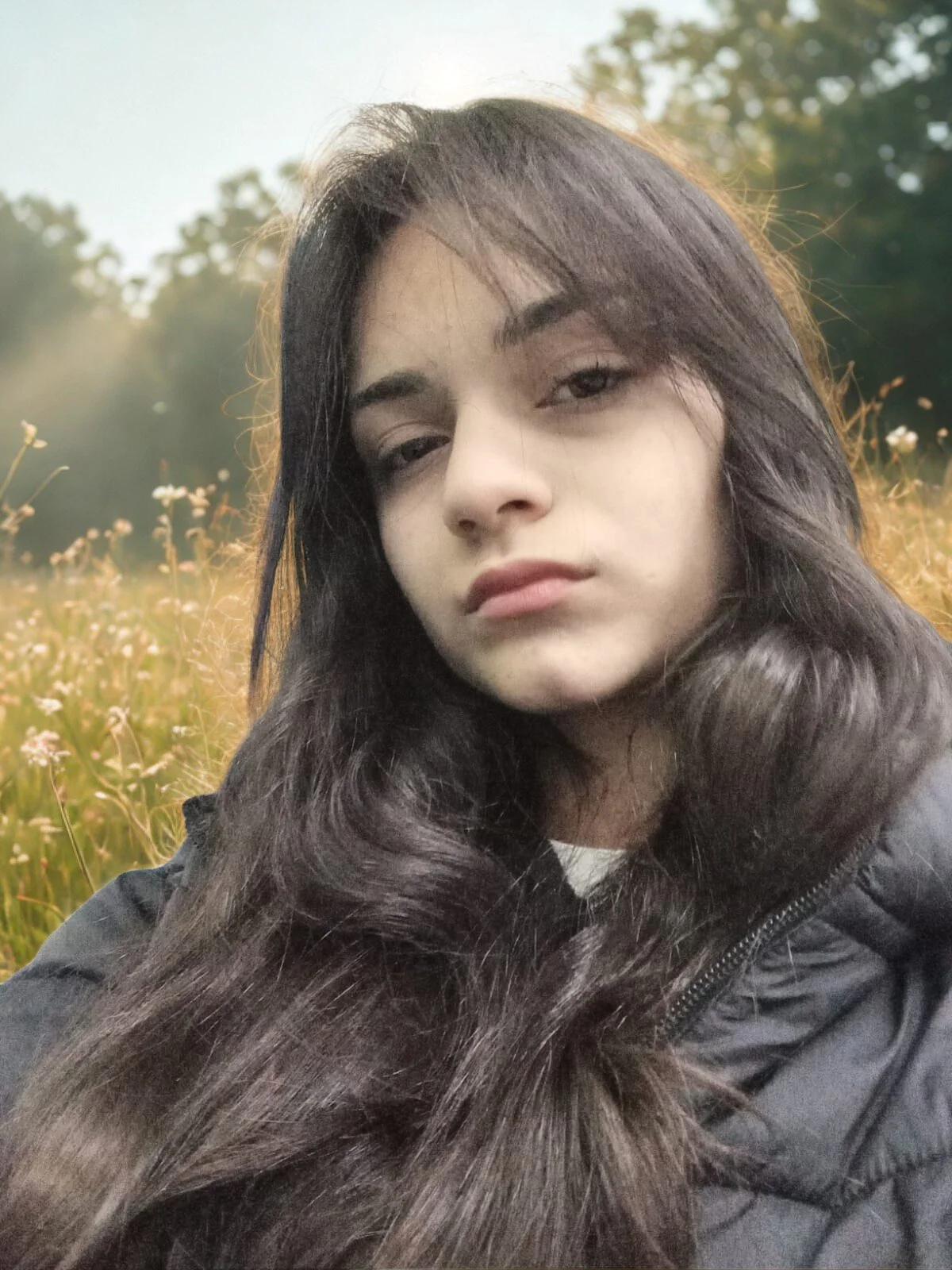 Kadın, açık alanda, çiçeklerle dolu bir tarlada, rüzgarda hafifçe savrulan uzun siyah saçlarıyla, gri bir mont giymiş ve hafifçe yana eğilmiş pozda.