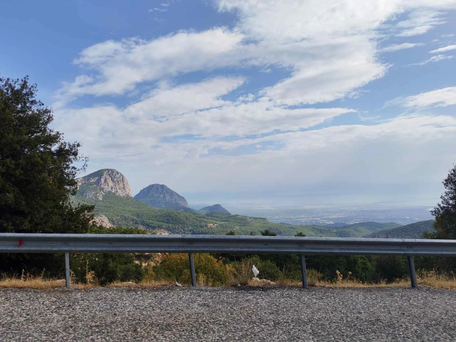 Dağlar ve ormanlar ile geniş bir manzara, ön planda metal bir bariyer ve yol yer almakta.