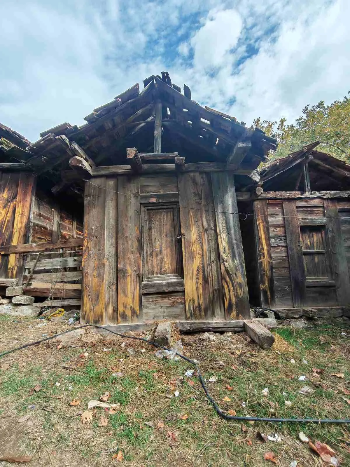 Eski ve harabe olmuş ahşap bir ev, çatısı ve duvarları ciddi şekilde hasar görmüş, dış cepheli birkaç pencere ve kapı var, zeminde yaprak ve atıklar bulunuyor, gökyüzünde bulutlar ve mavi gökyüzü görülüyor.