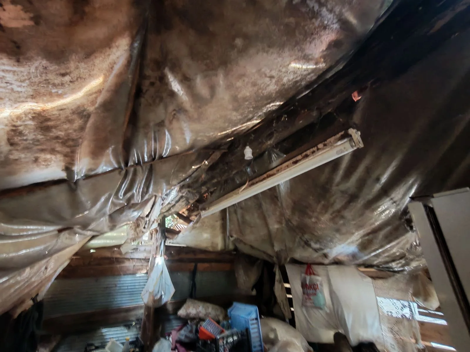 Tavana yığılmış ve plastik malzemelerle kaplanmış ıslak ve bozuk alan, çeşitli eşyalar ve ambalajlar da görülüyor