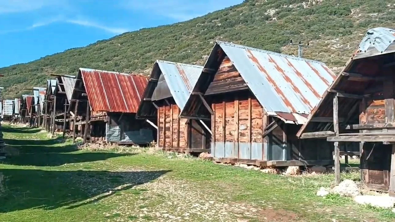 Dağ yamacında sıralanmış eski ahşap ve metal çatılı küçük evler, yeşil çimenlerle çevrili, gökyüzü açık ve mavi.