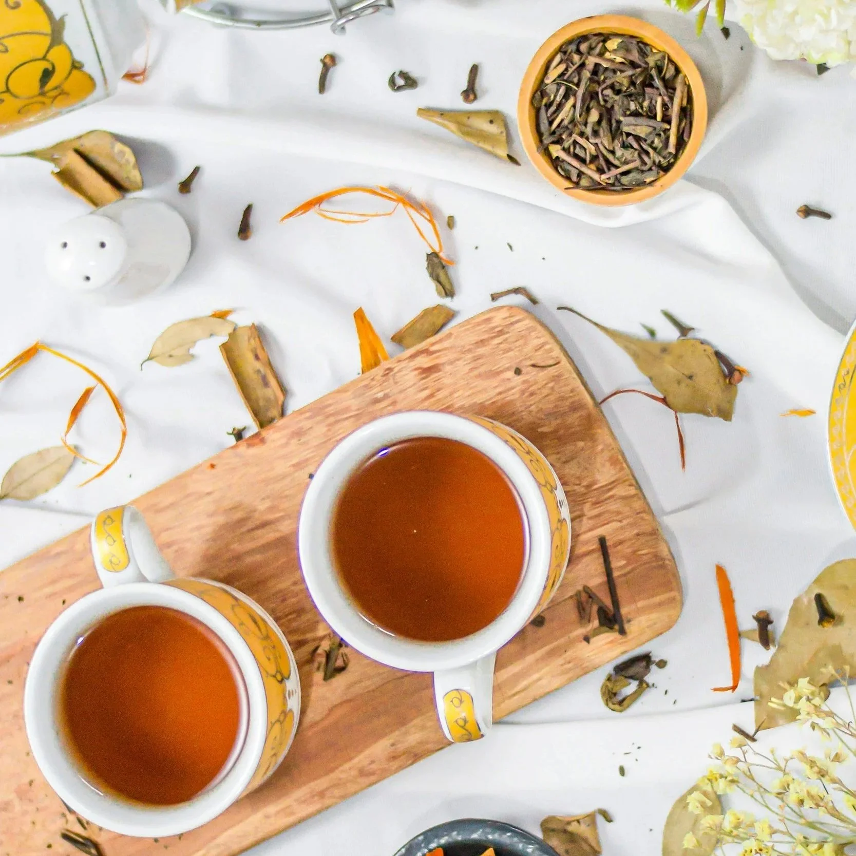 Two cups of tea on a wooden board surrounded by scattered dried leaves, a small bowl of loose tea leaves, a salt shaker, and flower petals on a white tablecloth.