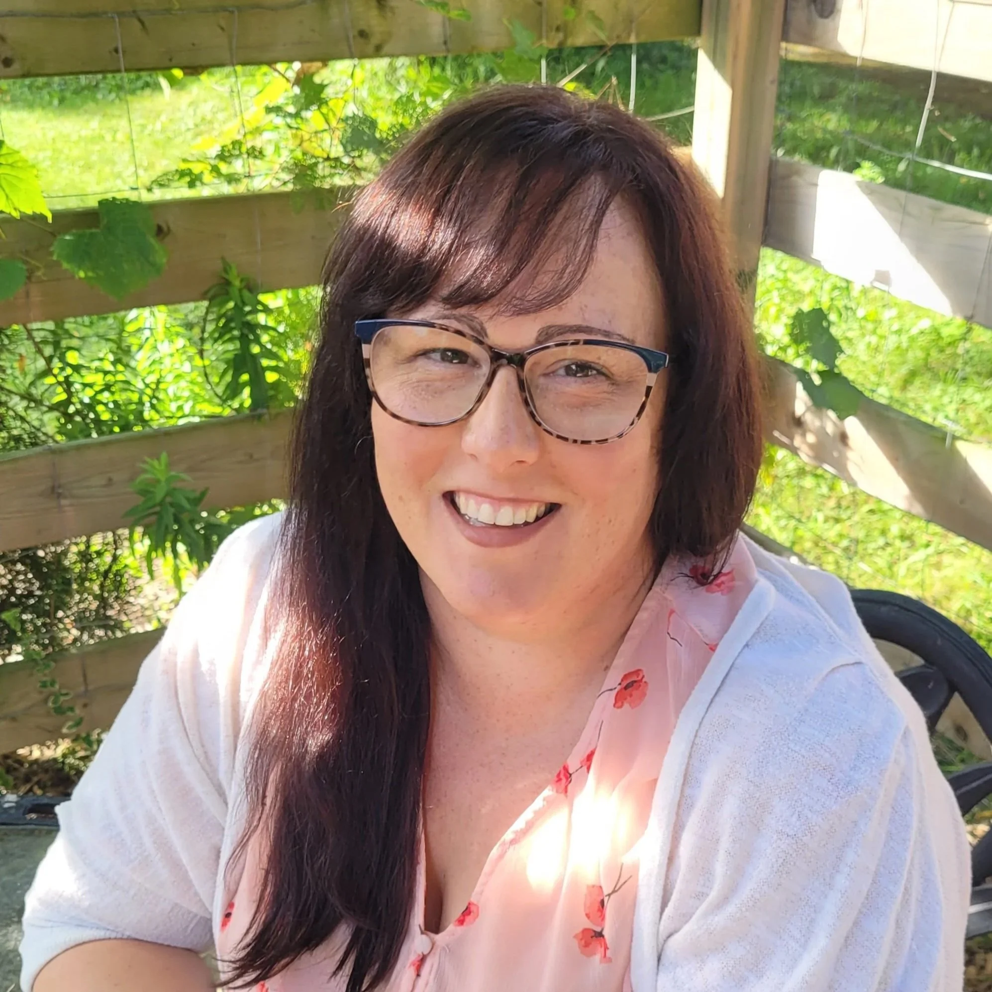 A woman with long dark hair and glasses smiling at a camera outdoors near a wooden fence with green foliage in the background.