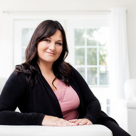 A woman with dark brown hair sitting on a white couch in a bright room with large windows.