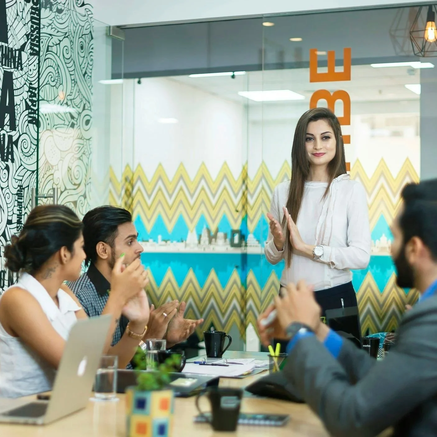 A woman standing in front of a glass wall, speaking to a group of four people seated at a table in a modern office conference room.