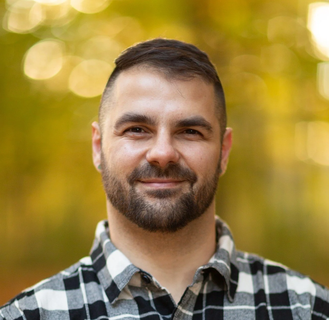 A smiling man with a beard and short dark hair, wearing a black and white checkered shirt, outdoors with a blurred background of trees and sunlight.