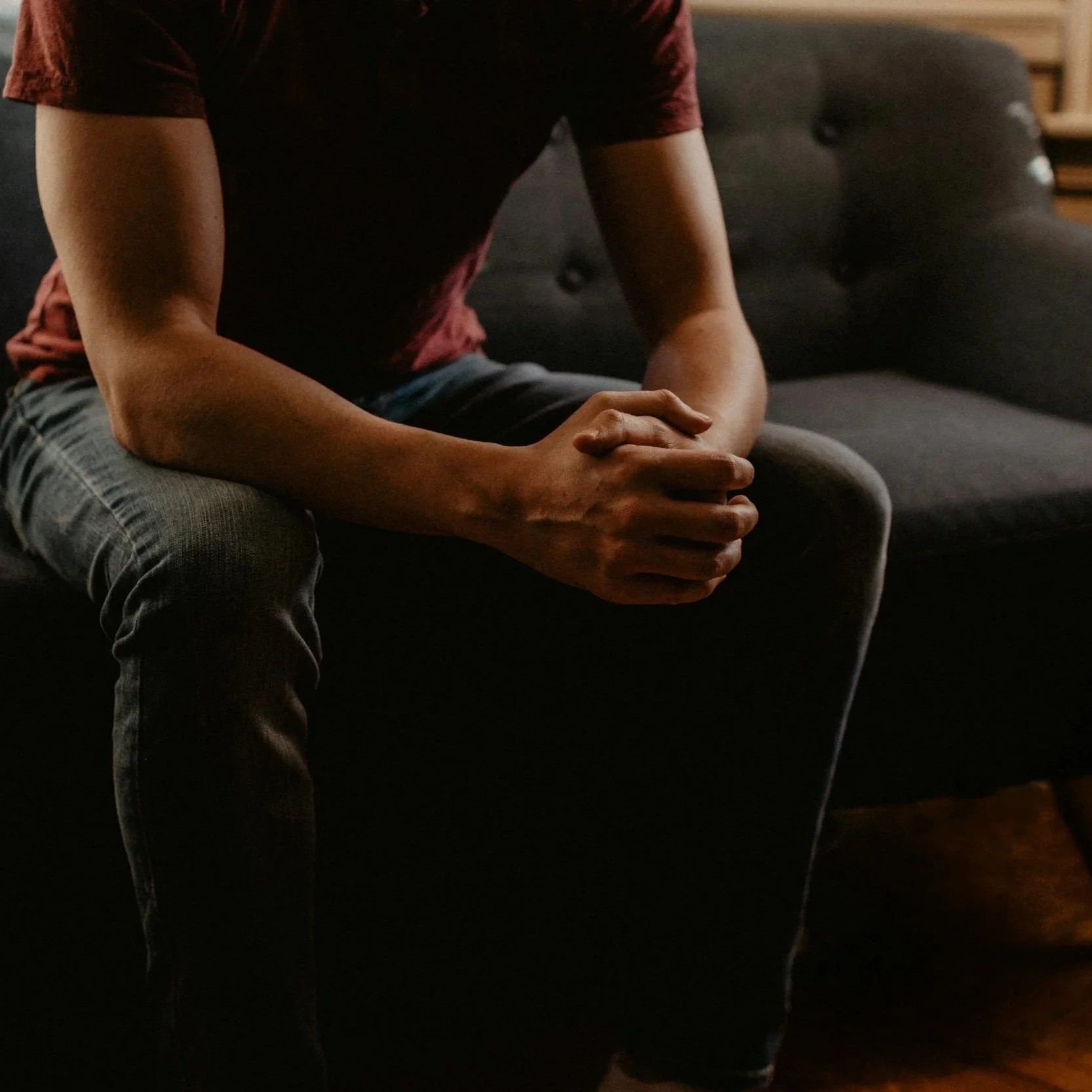 A person sitting on a black couch with hands clasped, wearing a red shirt and dark jeans, in a dimly lit room.