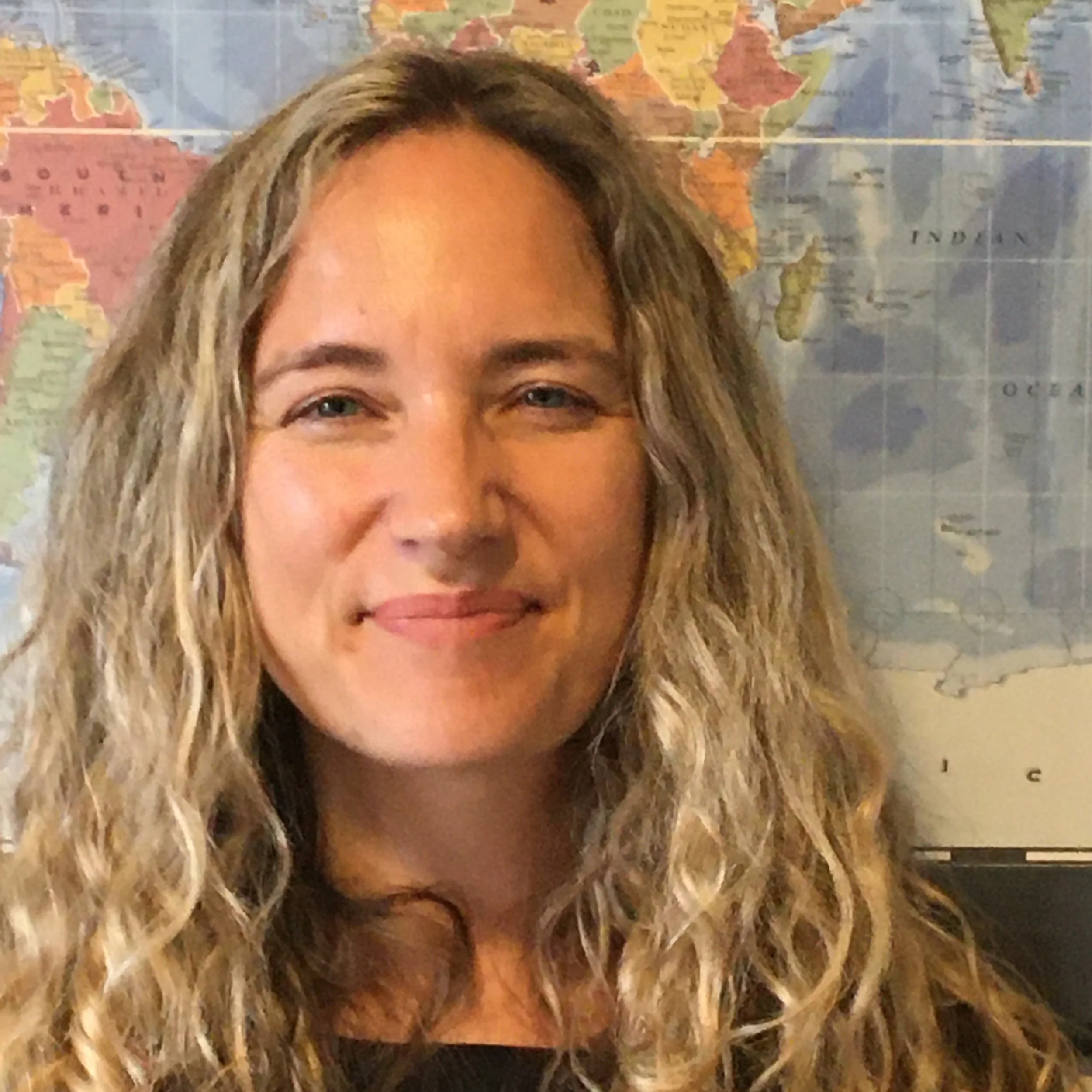 A woman with long, curly blonde hair smiling in front of a world map.