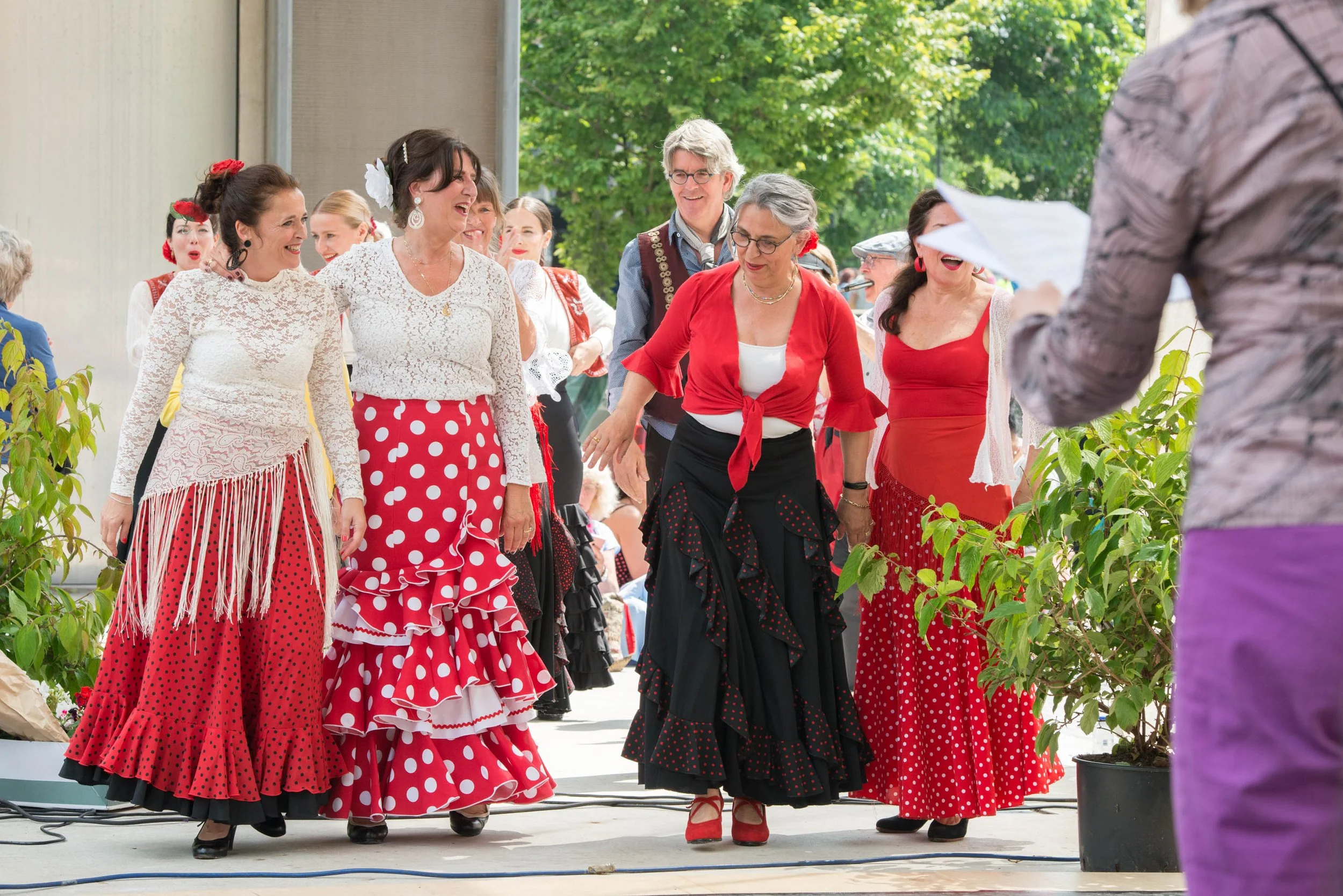 Group of women dancing and singing in traditional flamenco dresses, with an audience and greenery in the background.