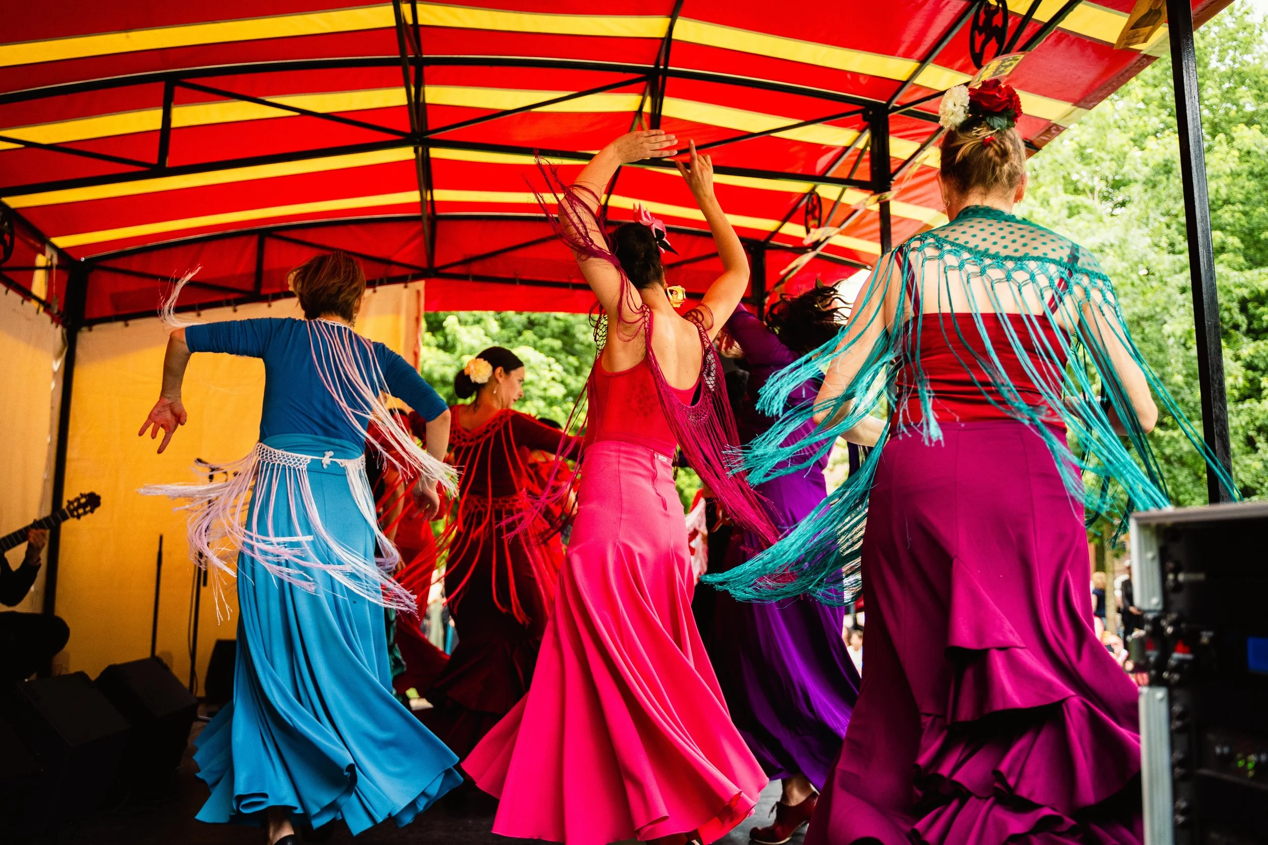 Women dancing in colorful flamenco dresses and shawls on an outdoor stage with a red and yellow striped canopy.