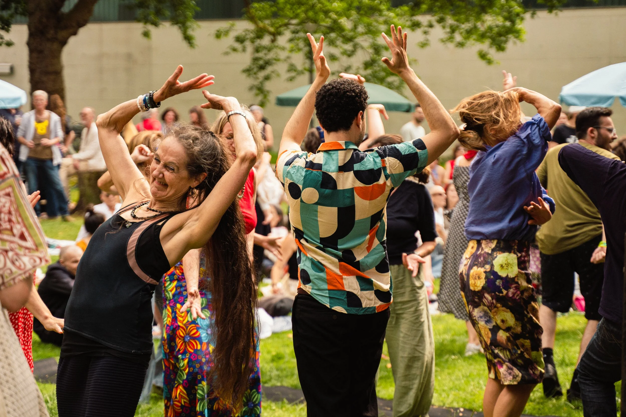 People dancing outdoors at a festival or gathering on a grassy area, with trees and umbrellas in the background.