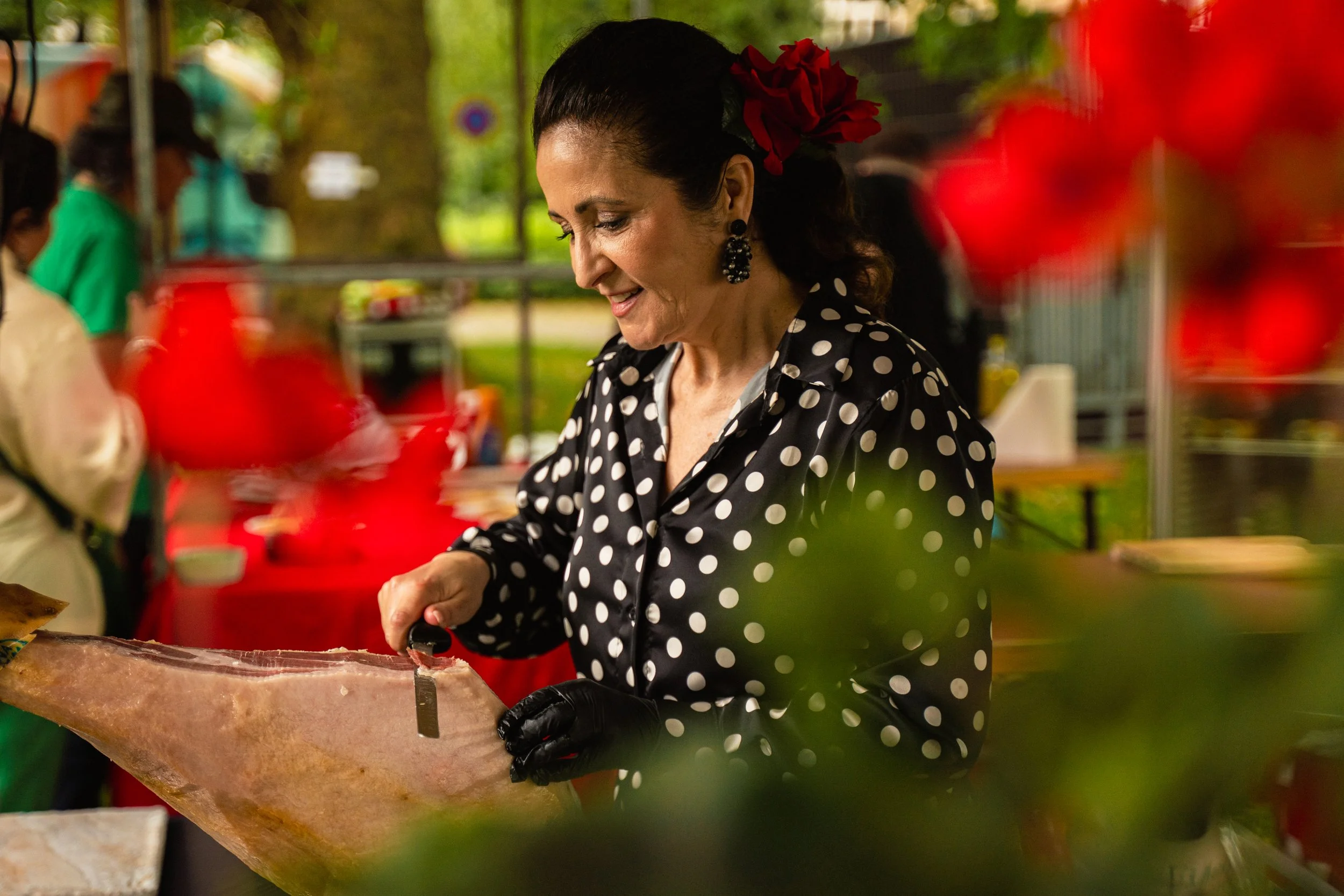 A woman slicing a large block of cured meat at an outdoor market, wearing a black dress with white polka dots and black gloves.