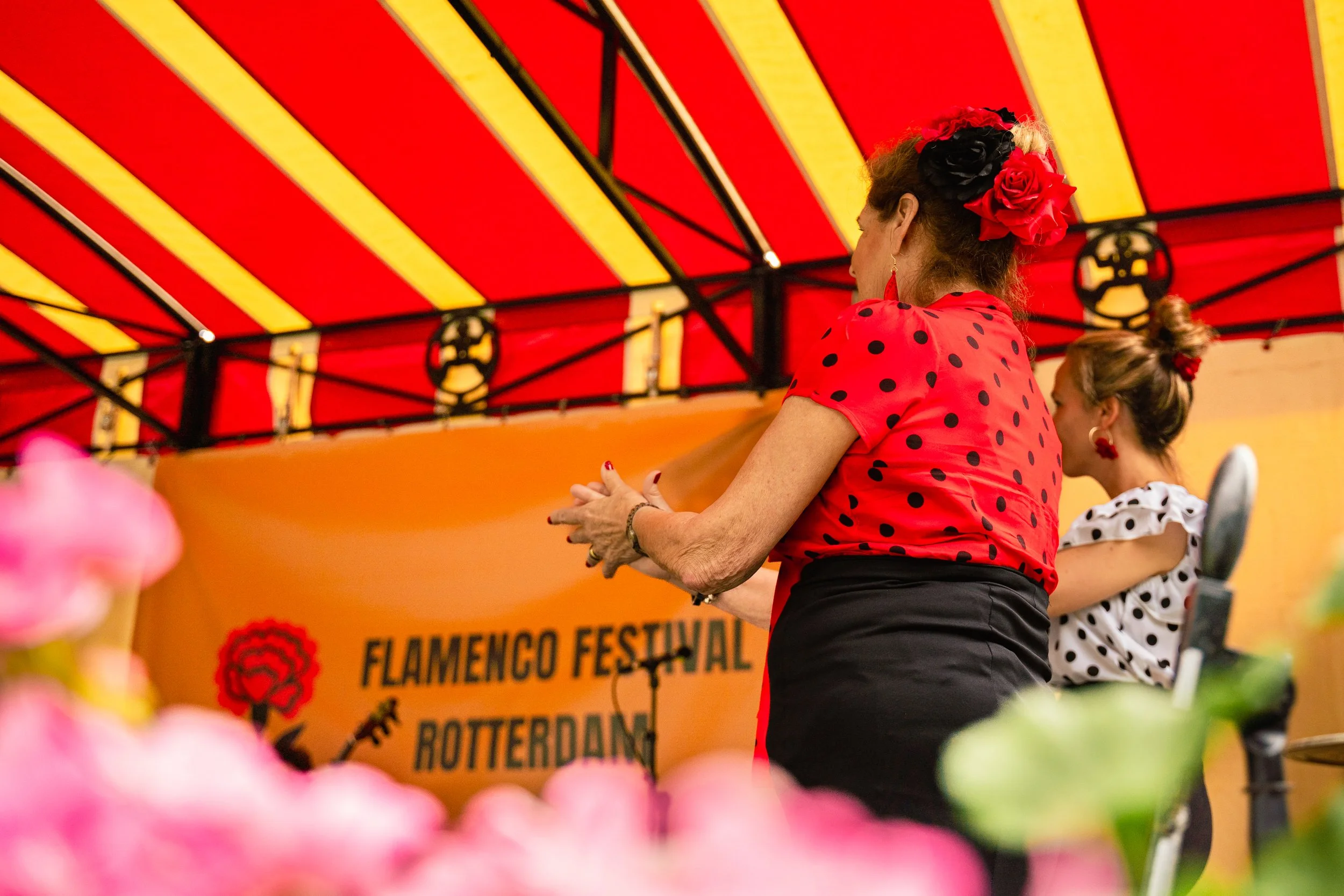Women in traditional flamenco attire on stage at a flamenco festival in Rotterdam, with flowers in the foreground and a colorful striped canopy overhead.