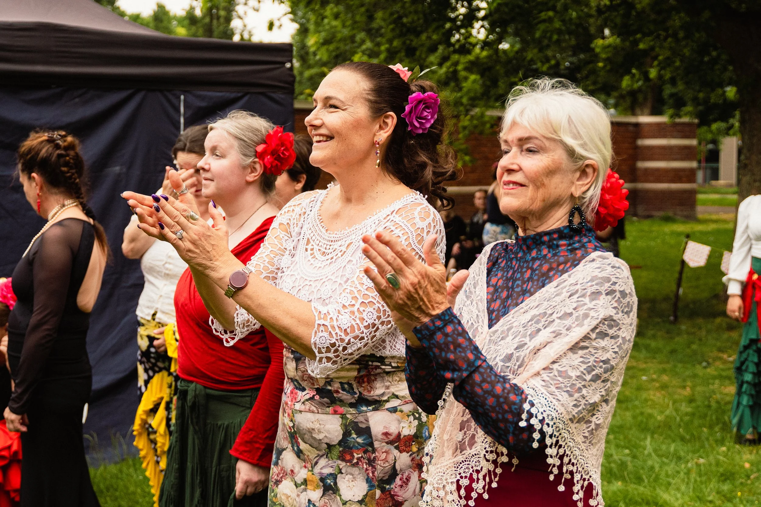 Group of women at an outdoor event, dressed in colorful, elegant clothing, clapping and smiling, with trees and a tent in the background.