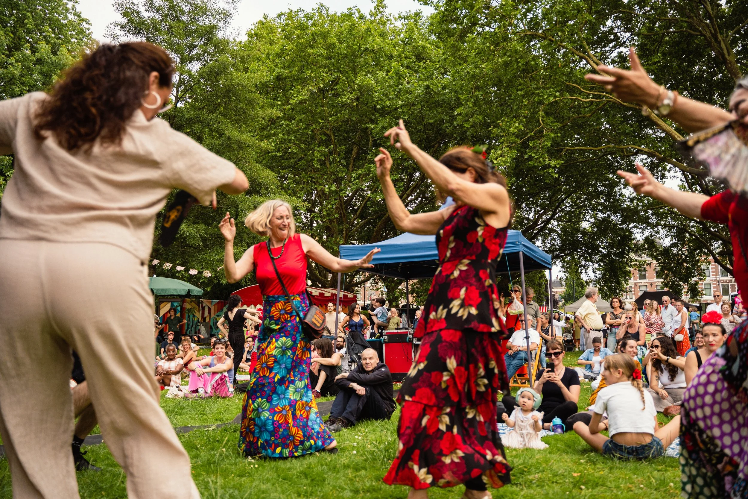 People dancing outdoors at a festival with trees in the background and a crowd watching.
