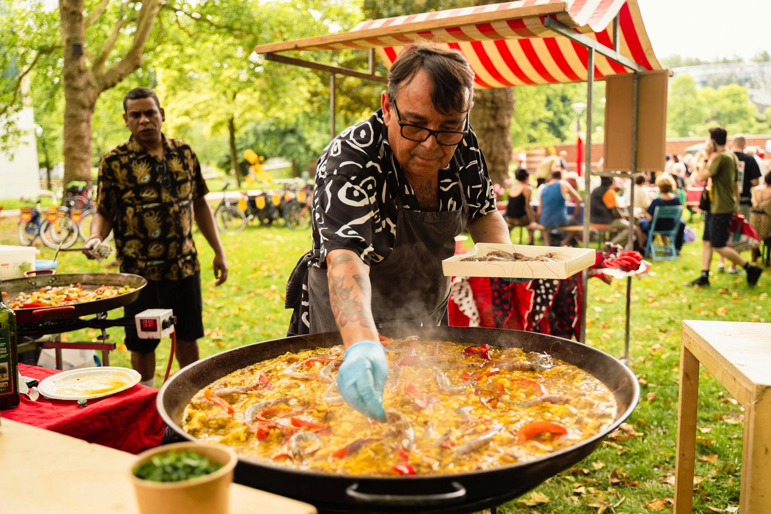 A man cooking seafood paella outdoors at a community event, with another man standing nearby and people sitting and walking in the background.