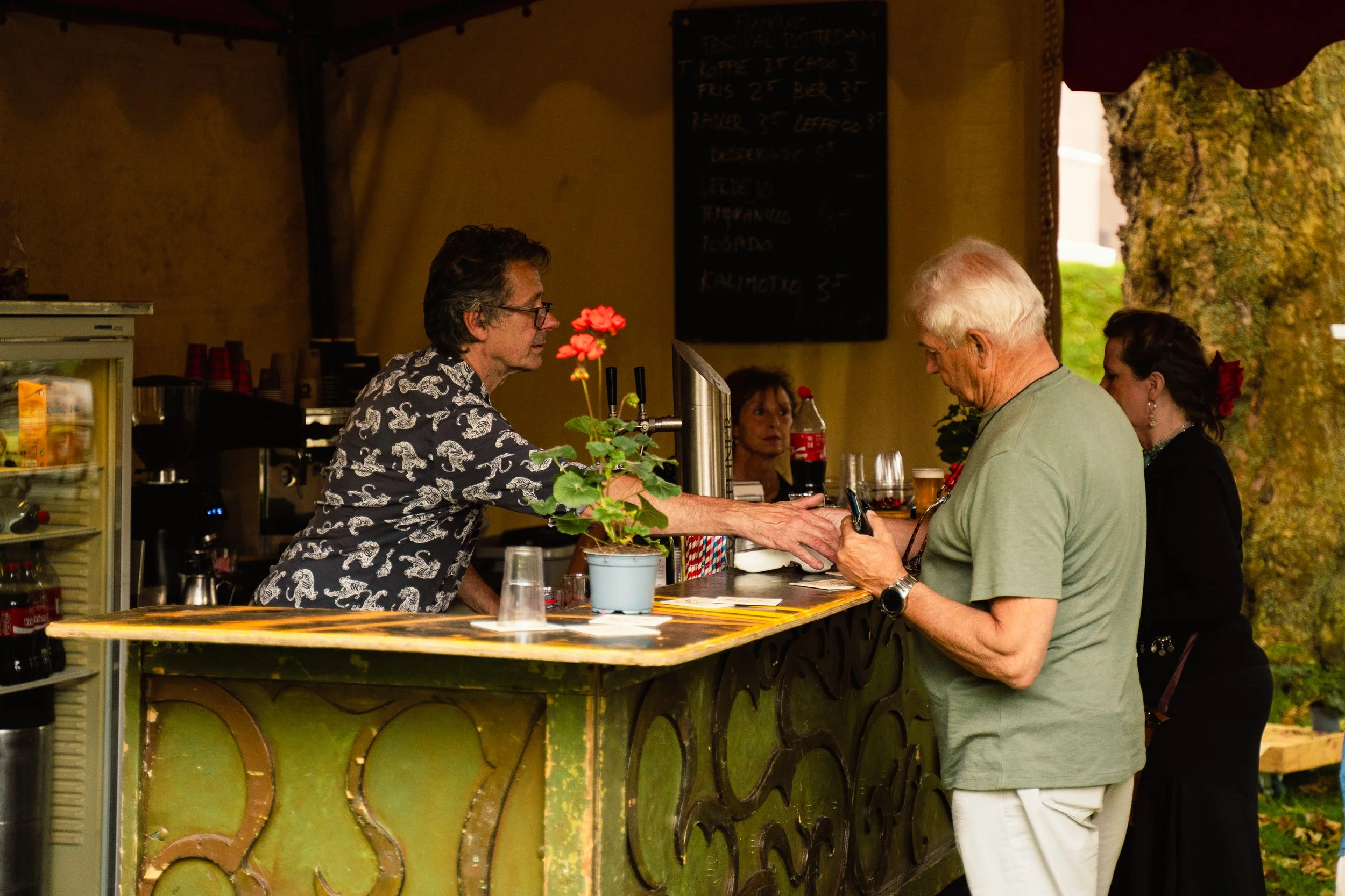 A bartender serving a customer at an outdoor bar with a decorative green counter, alongside a woman in black standing nearby. The scene is set outdoors with a tree and a blackboard menu in the background.