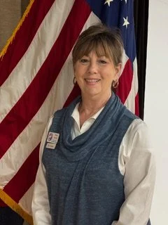 Professional headshot of Vice President Susan Jarrell wearing a white blouse and blue sleeveless sweater, standing in front of an American flag and smiling at the camera.
