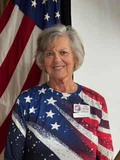 Professional headshot of Treasurer Victoria Hawkins wearing a patriotic American flag-themed top, standing in front of an American flag and smiling at the camera.