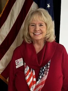 Professional headshot of Secretary Christine Roberts wearing a red top and scarf, standing in front of an American flag and smiling at the camera.