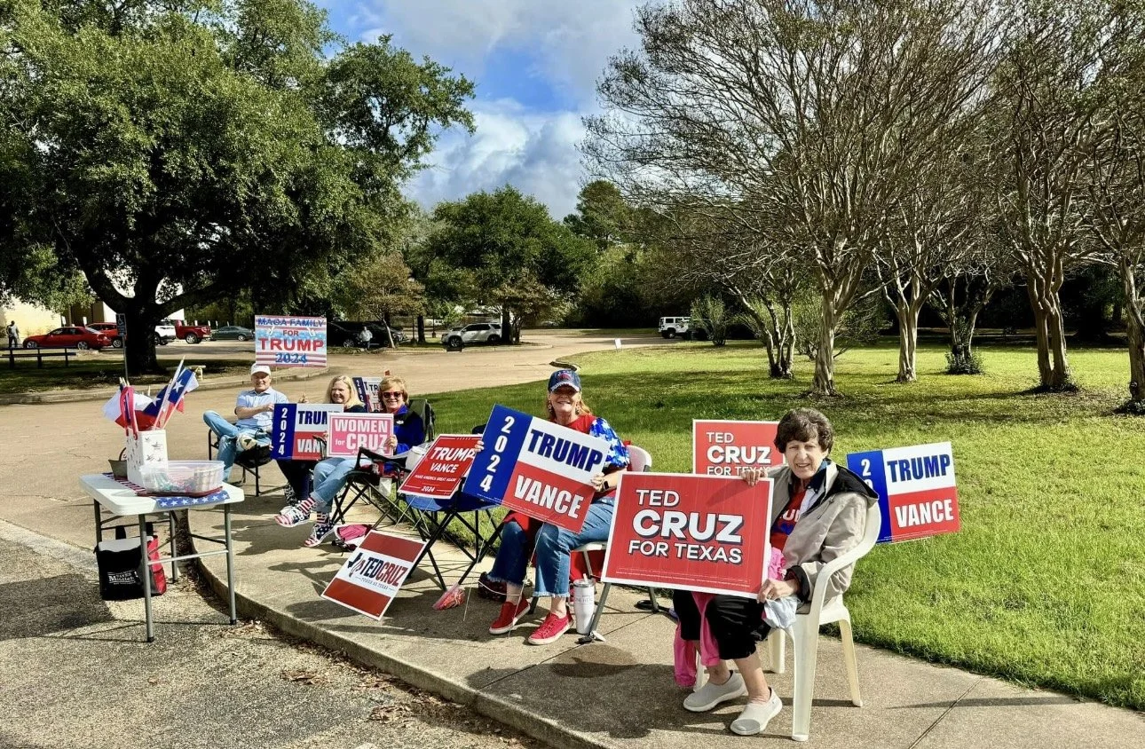 A group of women sitting outdoors holding campaign signs for Republican candidates including Ted Cruz, Donald Trump, and J.D. Vance. They are smiling and dressed in patriotic clothing, gathered near a grassy area with trees and blue skies overhead.