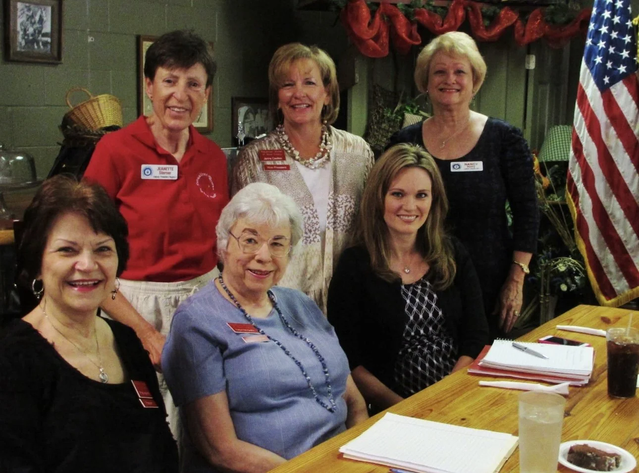 Six women seated and standing around a wooden table during a meeting, smiling at the camera. The setting includes notebooks, glasses of iced tea, and an American flag, showing members of the Wood County Republican Women gathered for discussion and fe