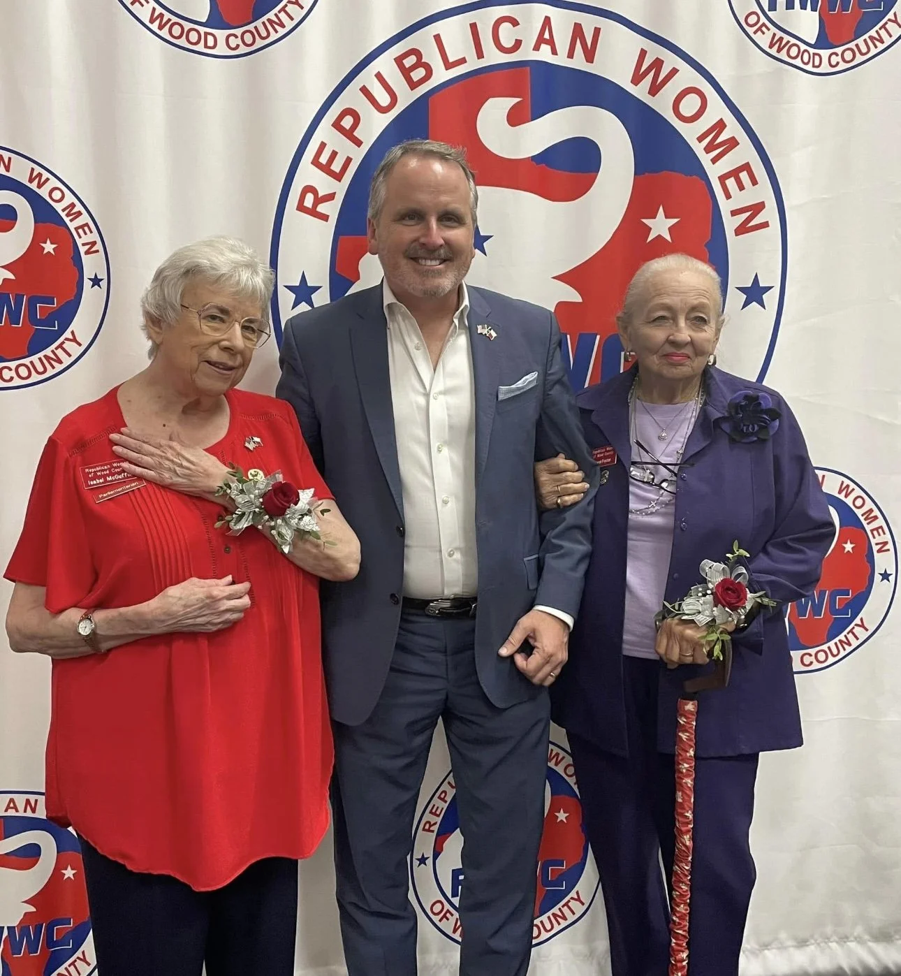 Three people standing in front of a backdrop with the logo of the Republican Women of Wood County, including two elderly women and a man. The women are wearing corsages with red roses, and the man is dressed in a blue suit.