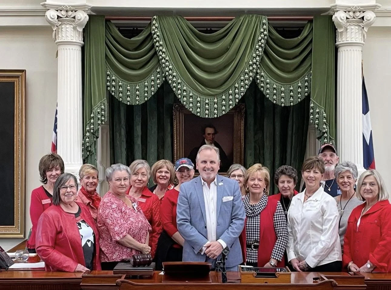 A group of women in red and white clothing stand behind a man in a light blue suit inside a formal government chamber with green drapes and Texas flags. They are smiling together for a group photo, representing the Wood County Republican Women during