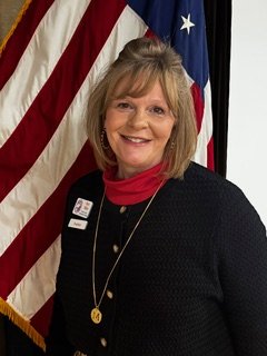 Professional headshot of a woman with shoulder-length blonde hair wearing a black top and red scarf, standing in front of an American flag and smiling at the camera.