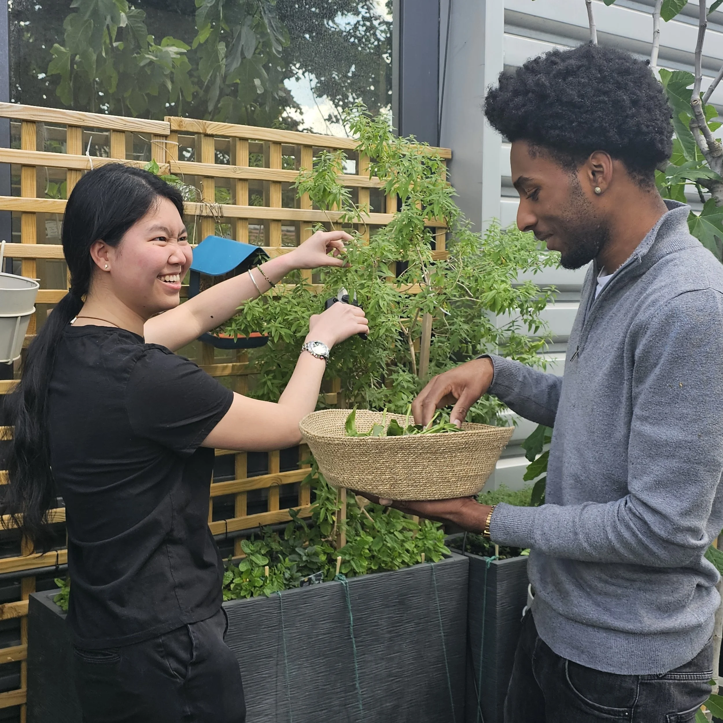 Deux personnes, une femme d'origine asiatique et un homme d'origine africaine, échangeant des plantes dans un environnement extérieur, entourées de verdure et de structures en bois.