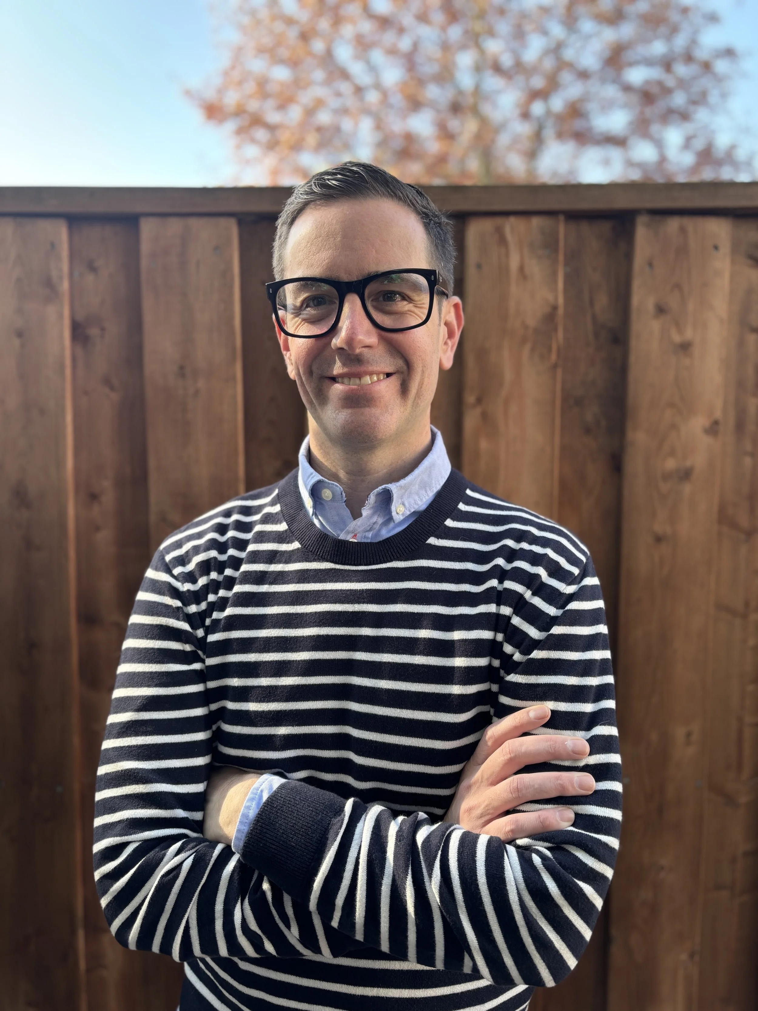 Image of Jason Magennis, high performance coach, standing outdoors in front of a wooden fence and autumn trees, smiling with arms crossed.