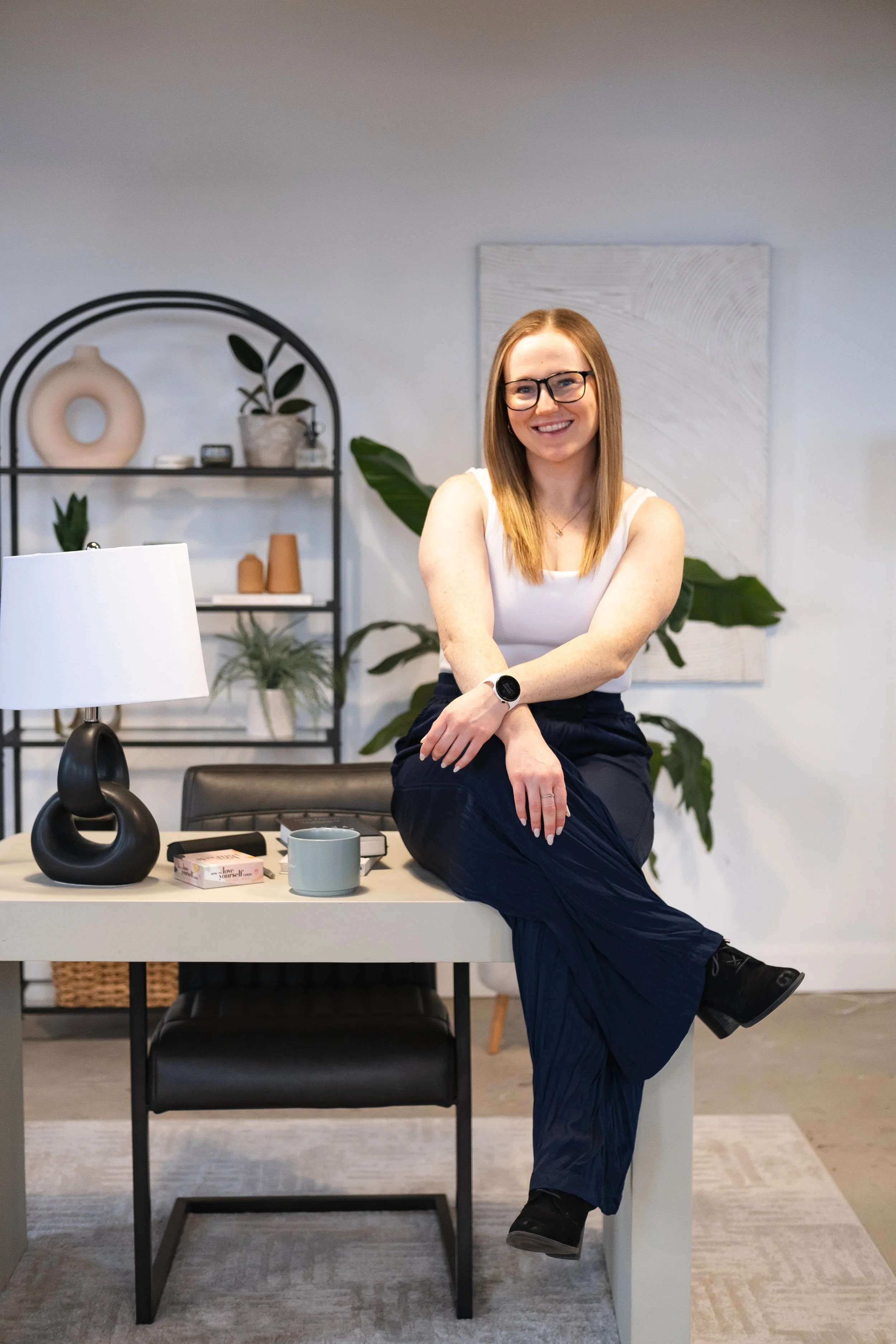 A strength coach for women with glasses and a white tank top sitting on a desk in an office, smiling.