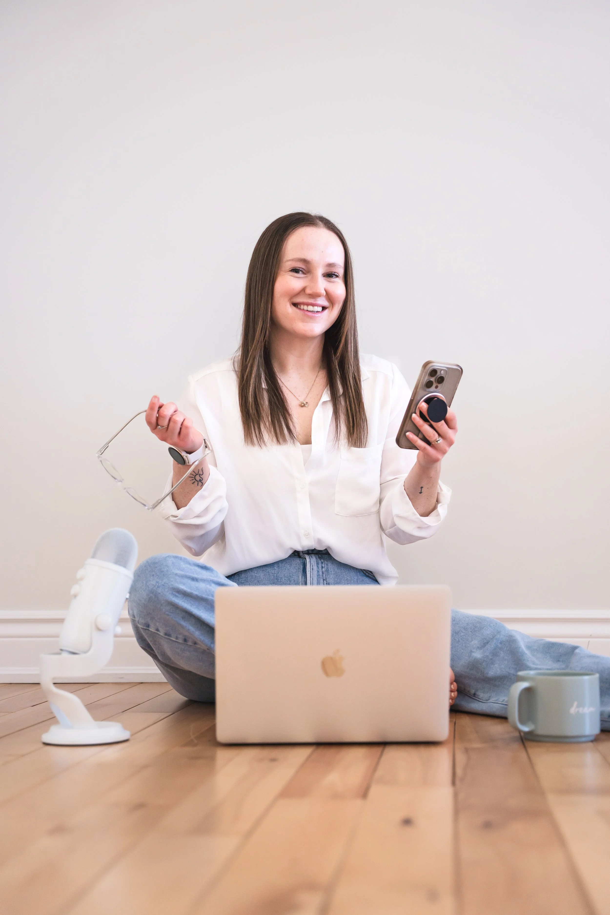 A young woman with long brown hair, sitting on a wooden floor, smiling while holding a smartphone with a pop socket. She is wearing a white blouse and blue jeans. There is a gray mug to her right, a white microphone on a stand to her left, and a closed silver MacBook in front of her.