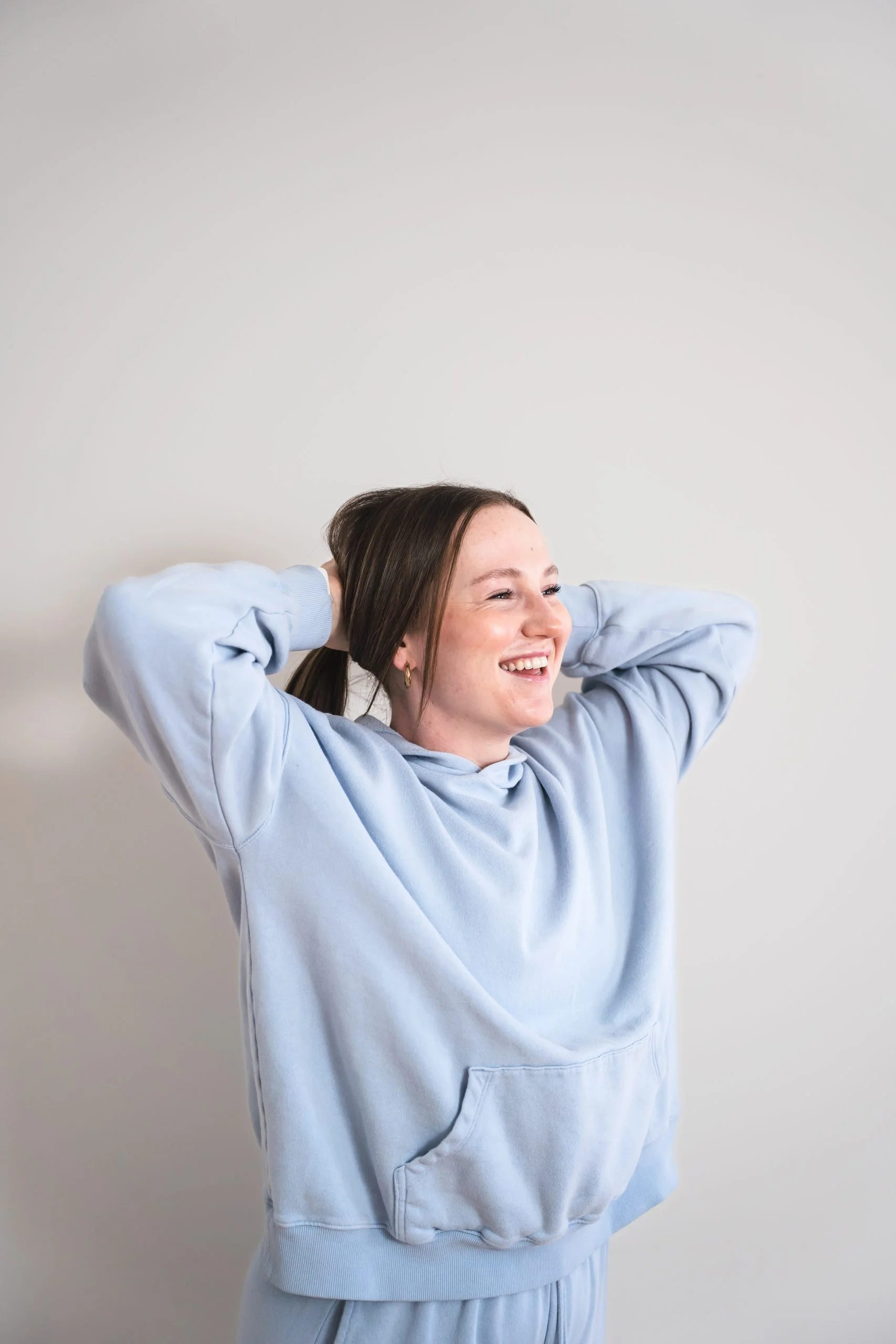 Confident women's strength coach in Brantford Ontario smiling with her hands behind her head with a light blue hoodie on.