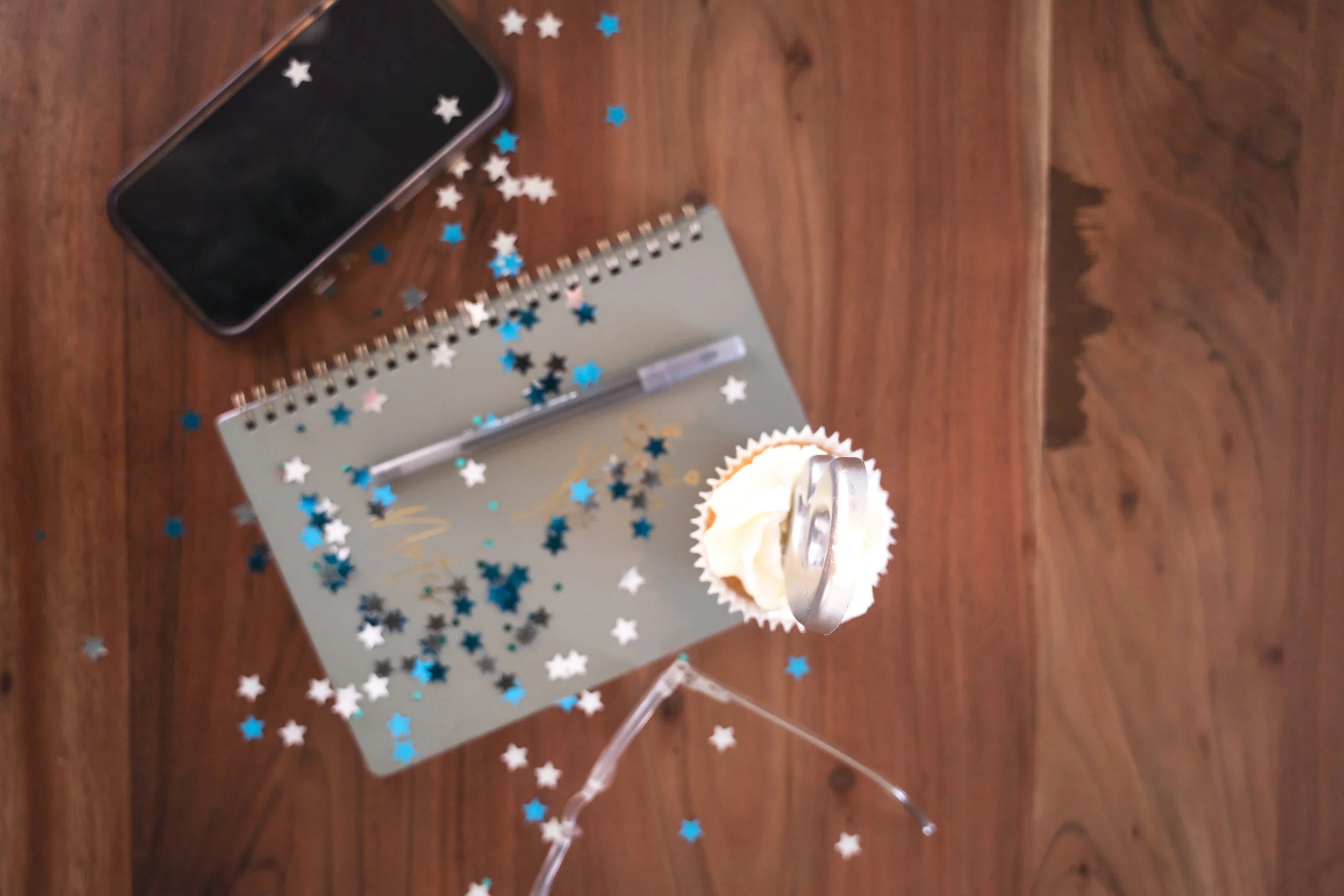 Overview of a personal trainer’s iPhone, notebook, pen, confetti, cupcake with white frosting and a pair of glasses on a table in Brantford.