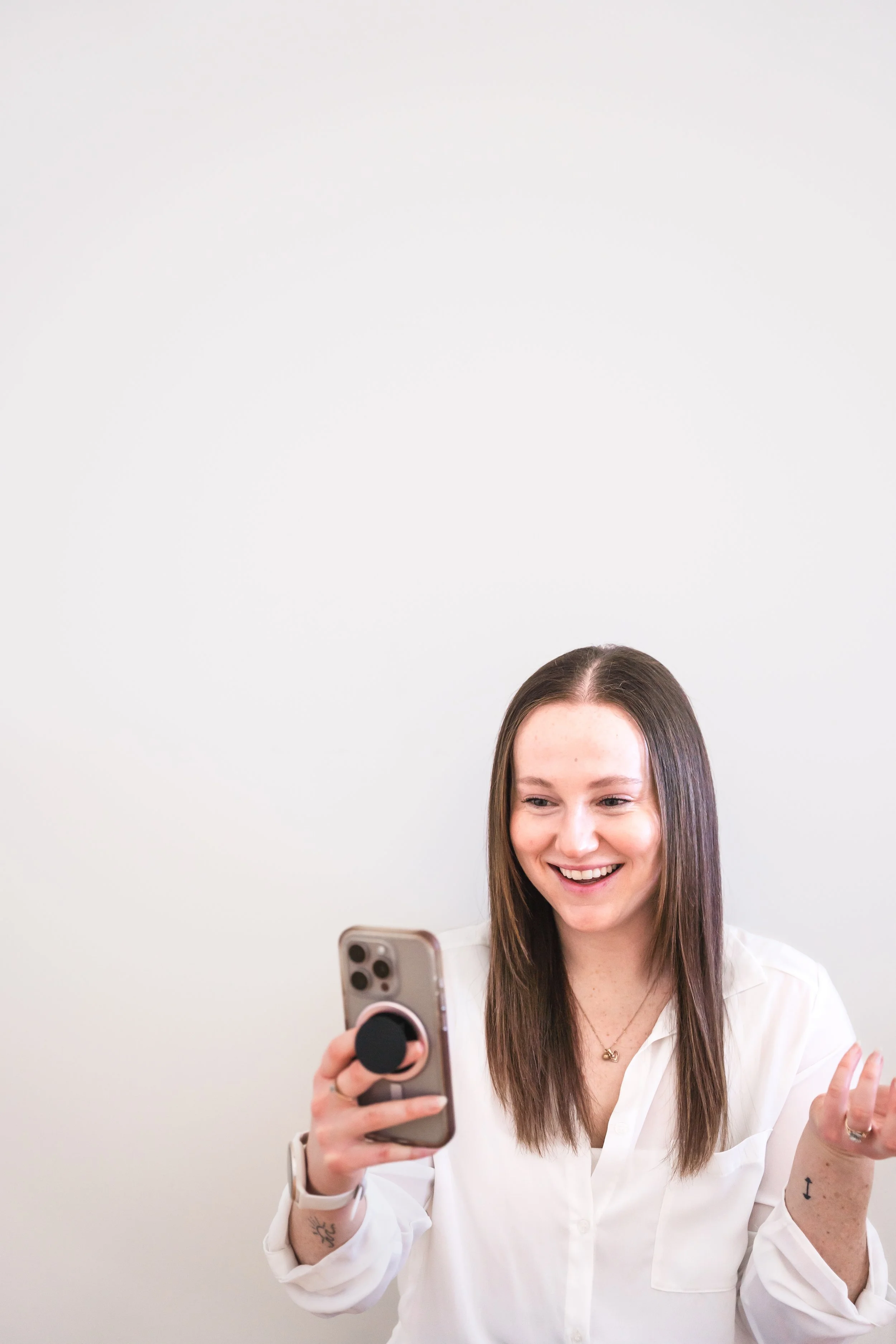 A young woman with long brown hair taking a selfie with her smartphone, smiling, wearing a white blouse, standing against a plain white wall.