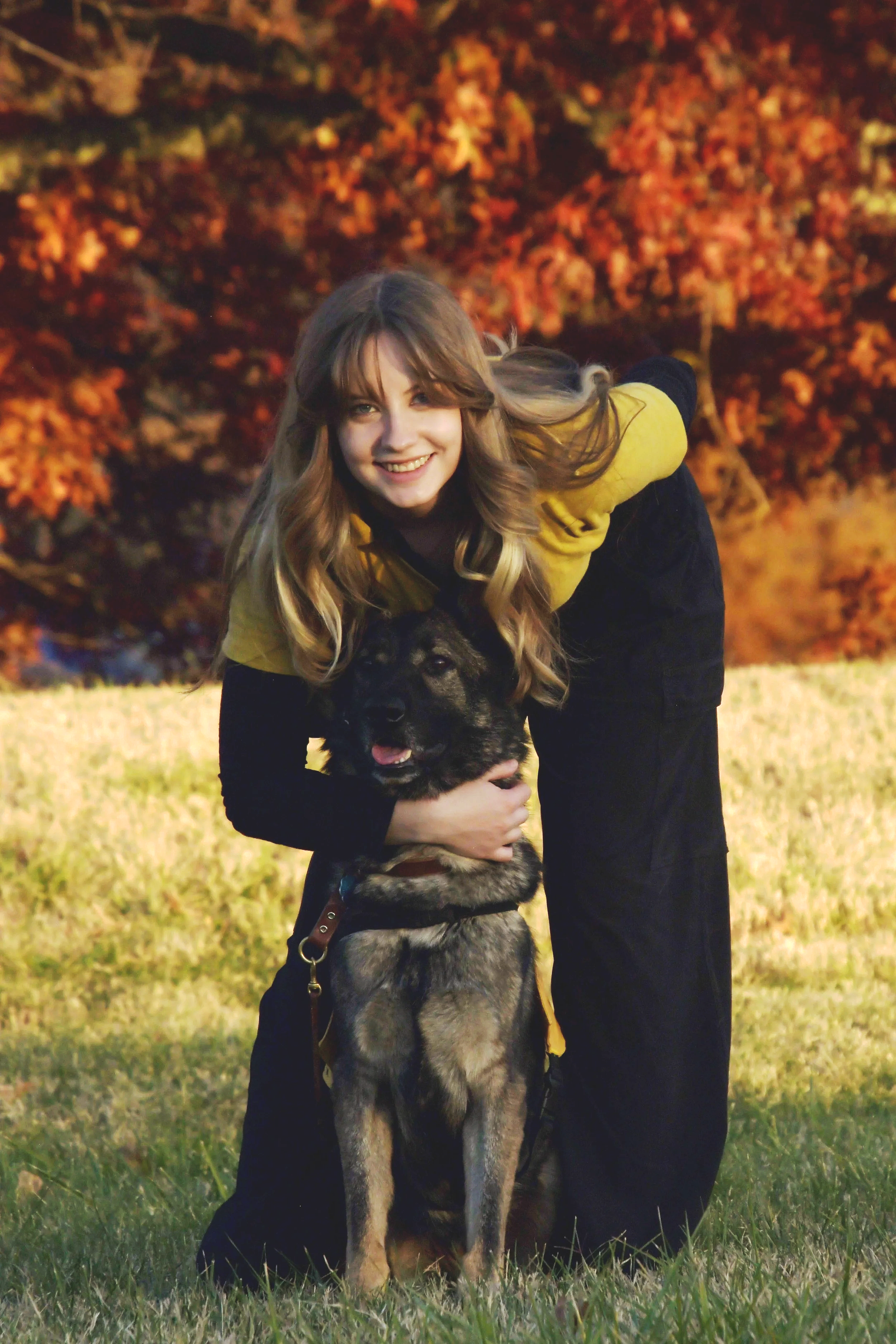 A young woman with long, wavy blonde hair smiling and crouching down while holding a black and tan German Shepherd puppy, outdoors on grassy ground with orange and red autumn trees in the background.